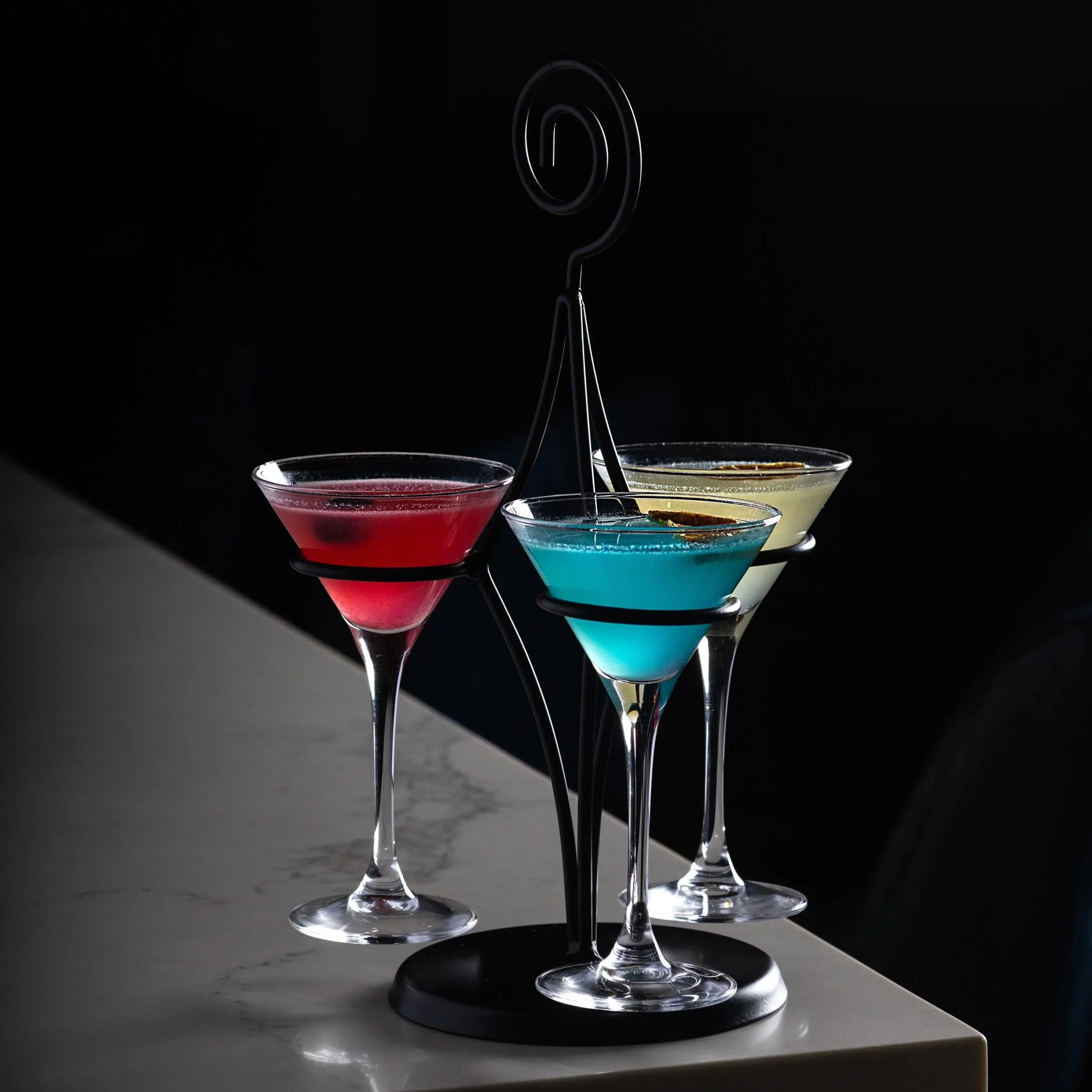 Three colourful Tiny Tini cocktails in martini glasses on a black metal stand, with a dark background and white marble countertop of Southside Shake's bar top.