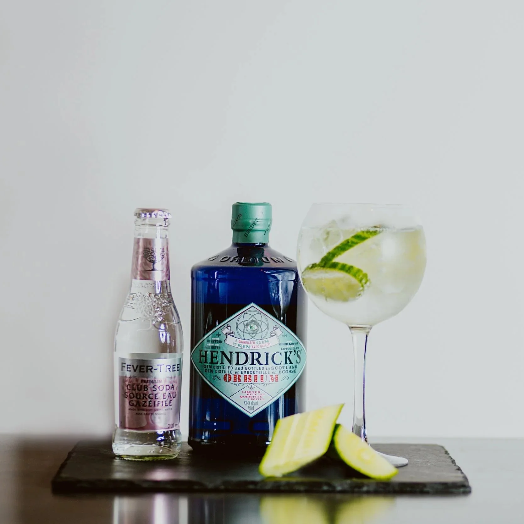 A bottle of Hendrick's gin, a bottle of Fever-Tree tonic water, and a glass of gin and tonic with lime and cucumber slices on a dark wooden tray against a plain white wall.