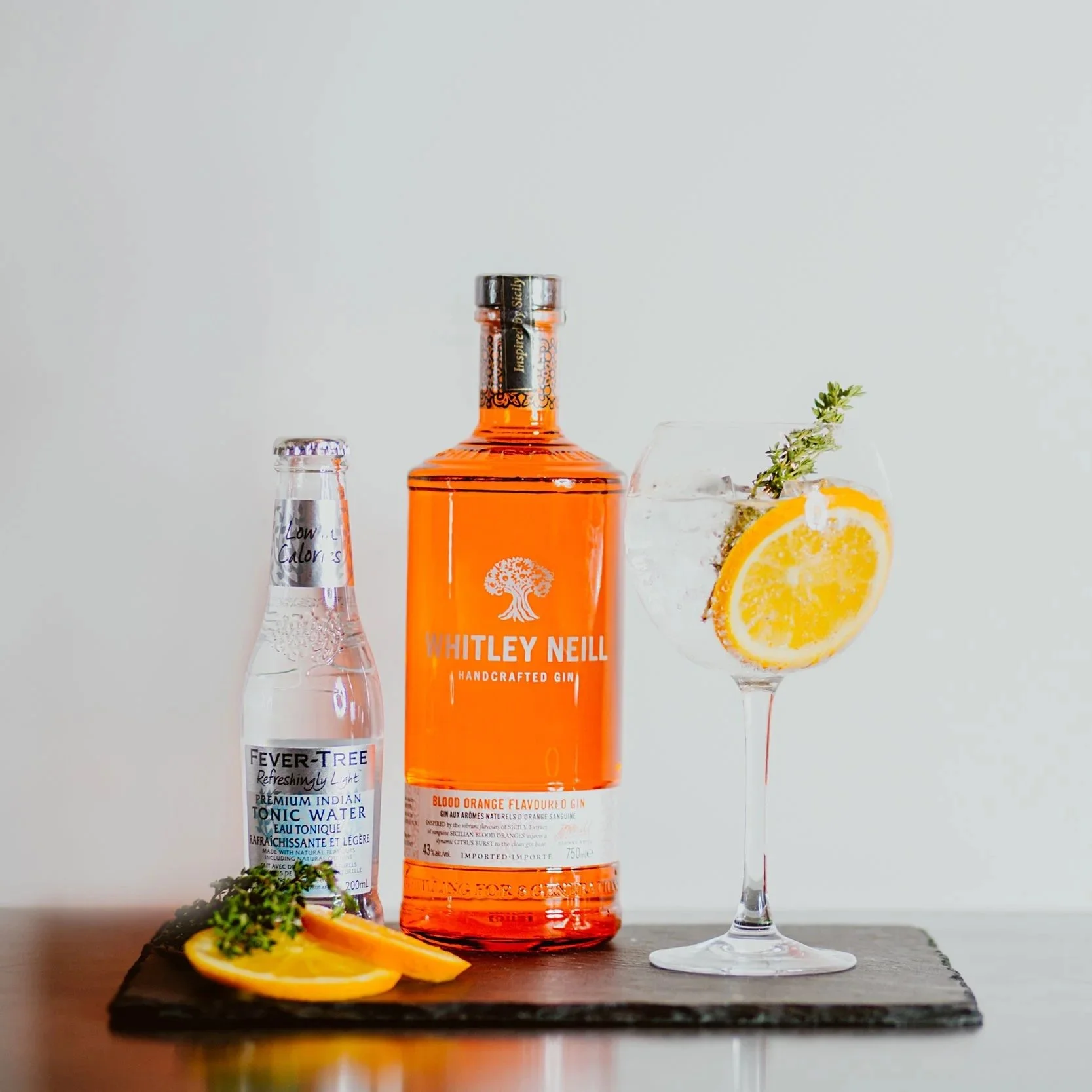 A bottle of Whitley Neill handcrafted gin, a bottle of Fever-Tree Indian tonic water, and a gin and tonic cocktail garnished with a lemon slice and a sprig of thyme on a black slate board.