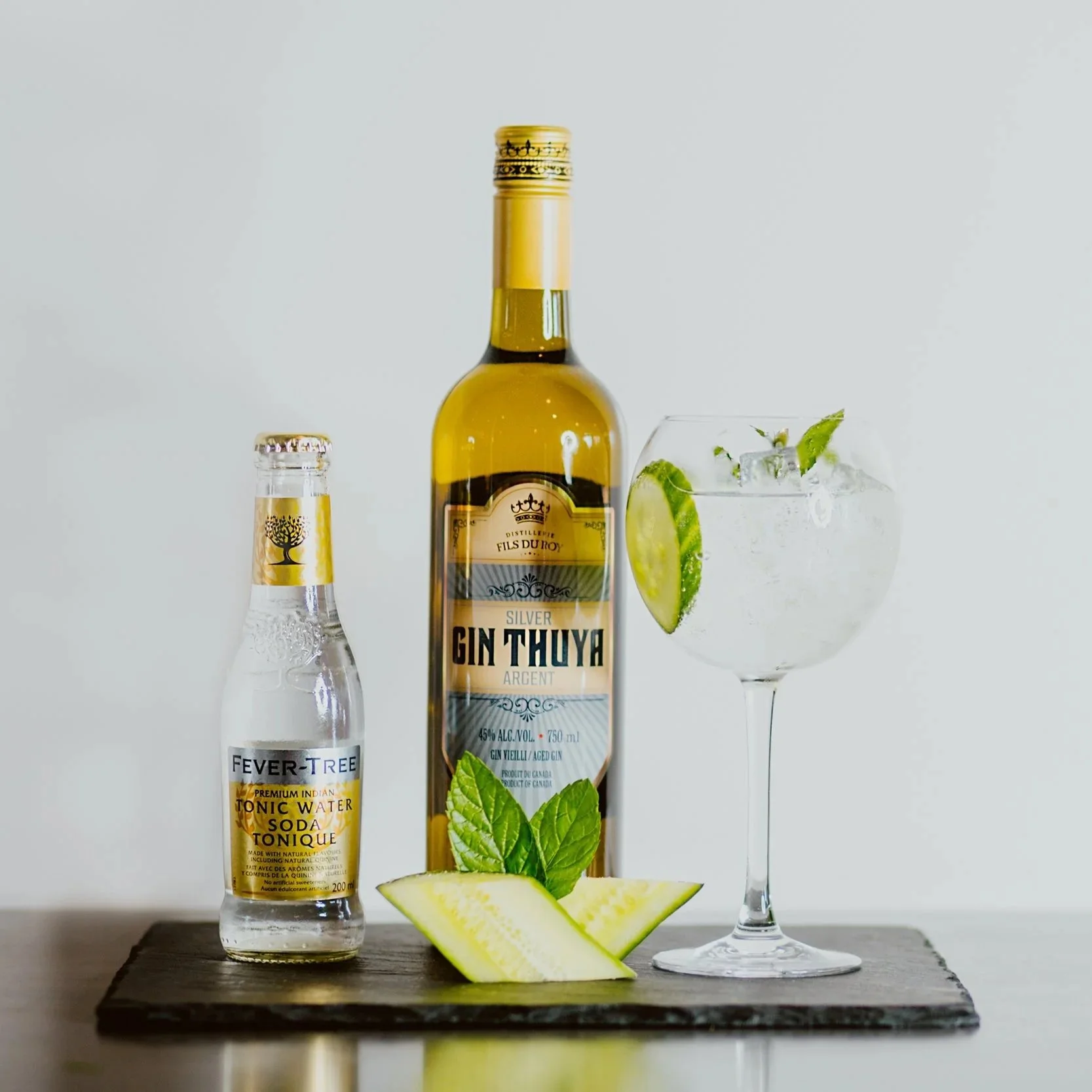 A bottle of Gin Thuyra, a small bottle of Fever-Tree tonic water, and a glass of gin and tonic garnished with lime and mint on a black tray with lime wedges and mint leaves.