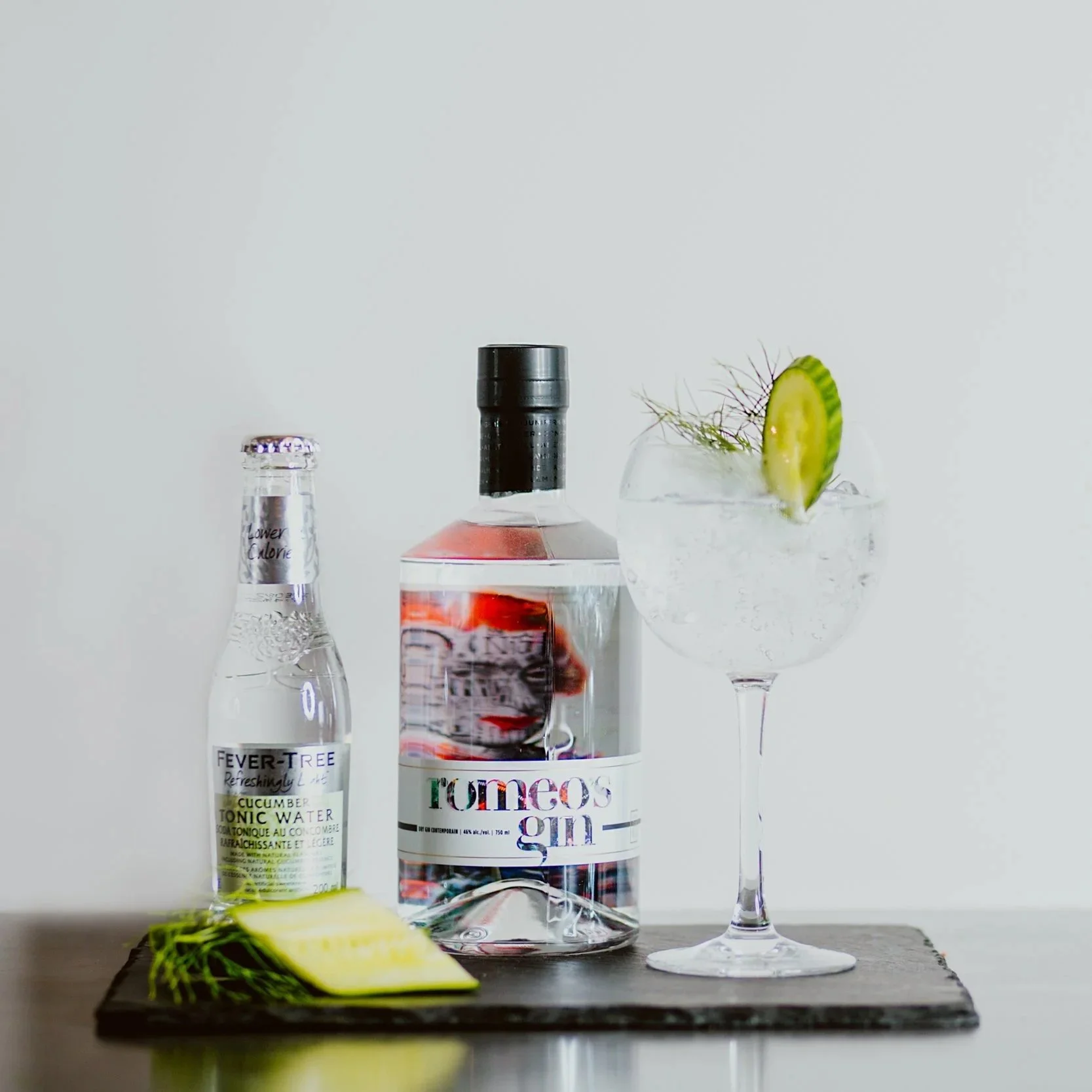 A bottle of rum, a bottle of tonic water, and a cocktail glass with a slice of cucumber and a sprig of herbs on a dark wooden surface against a plain white background.