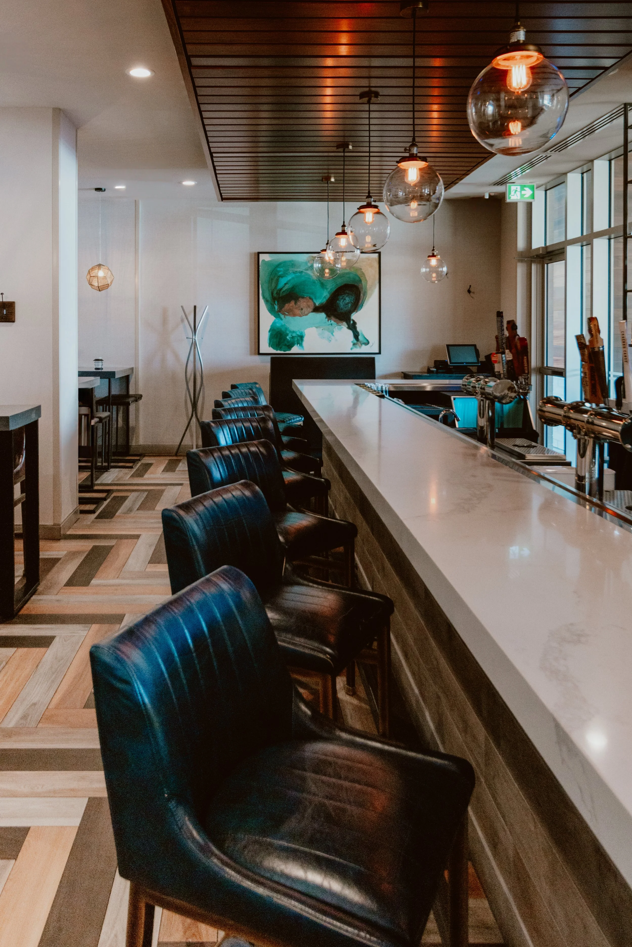 The modern bar interior of Southside Shake with navy leather chairs along a white marble bar counter, hanging glass globe pendant lights, wooden flooring, and sunlight coming through large windows.