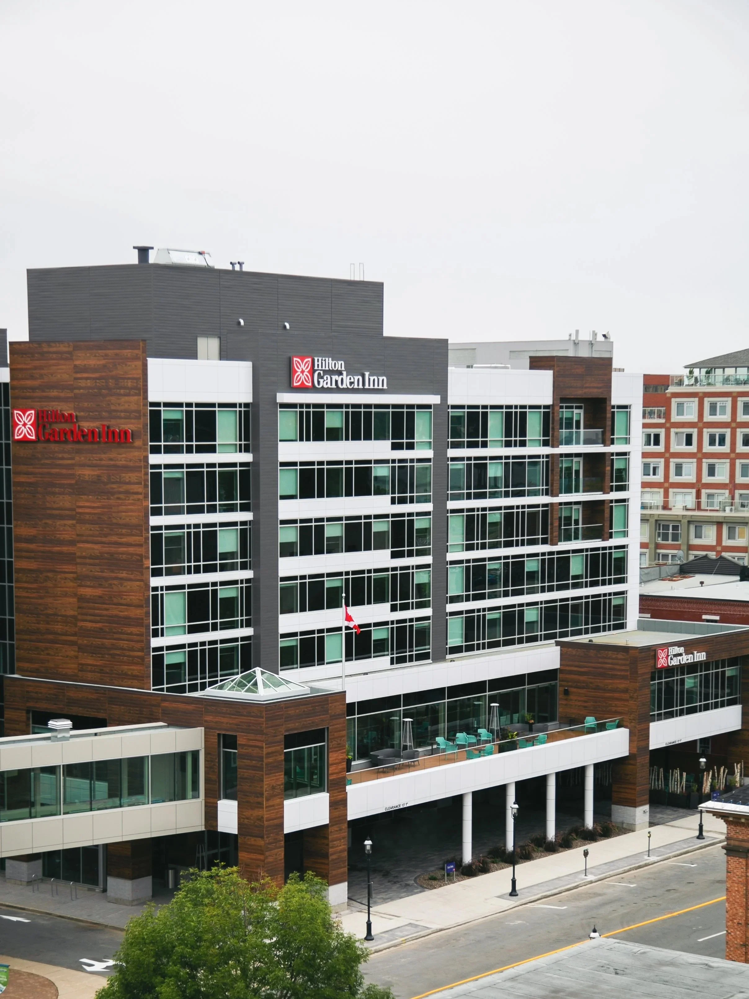 Modern multi-story Hilton Garden Inn Fredericton hotel with dark gray and wood exterior, red Hilton sign, and Canadian flag on balcony, in an urban setting of Fredericton's downtown.
