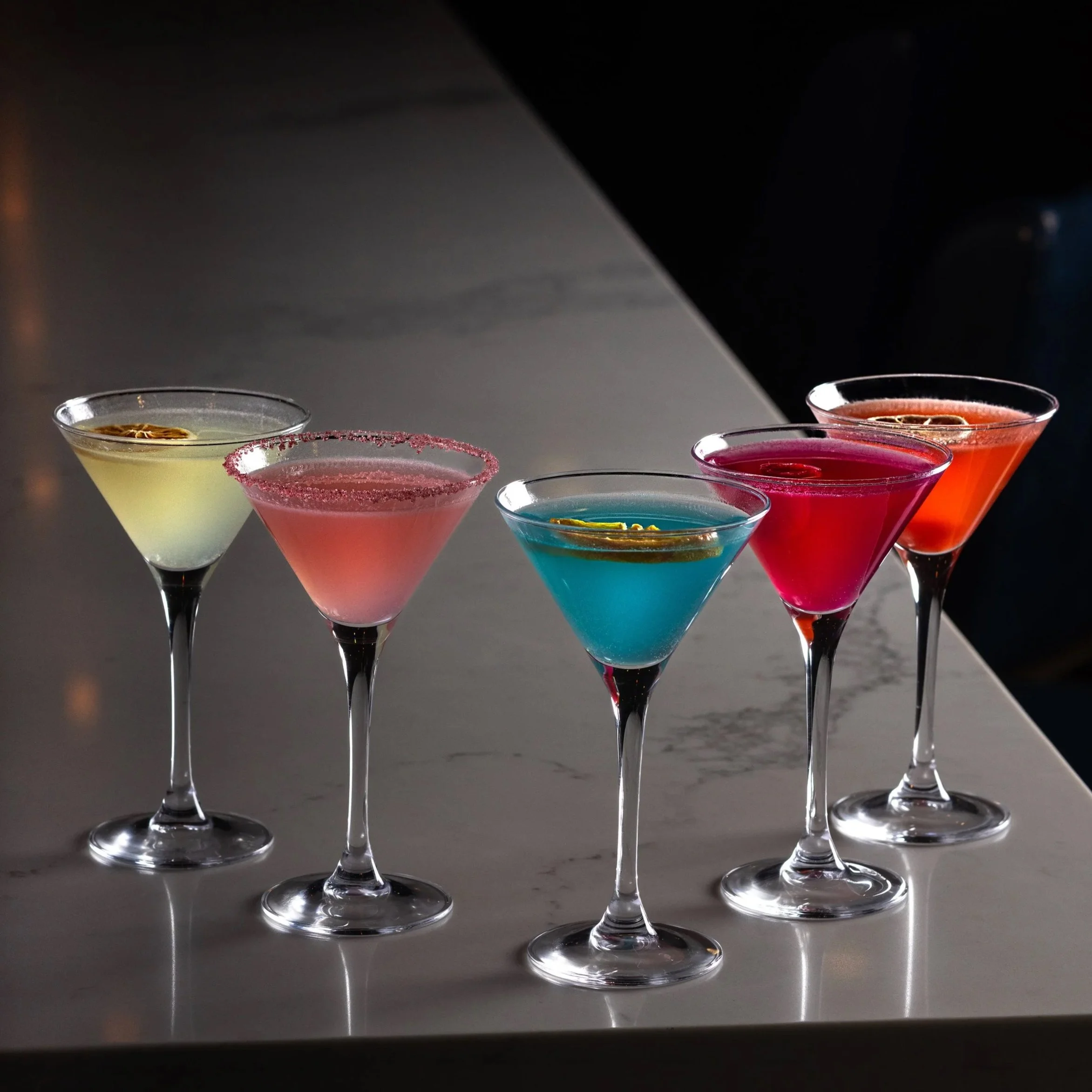 Five colourful Tiny Tini gin cocktails in martini glasses arranged on the marble bar counter of Southside Shake, each garnished with a slice of citrus.