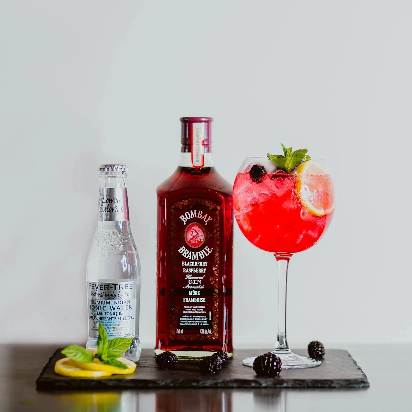 A bottle of Bombay Bramble blackberry raspberry gin, a bottle of Fever-Tree Indian tonic water, and a glass of pink cocktail with lemon slices, blackberries, and mint, all on a dark slate serving board with blackberry garnishes.
