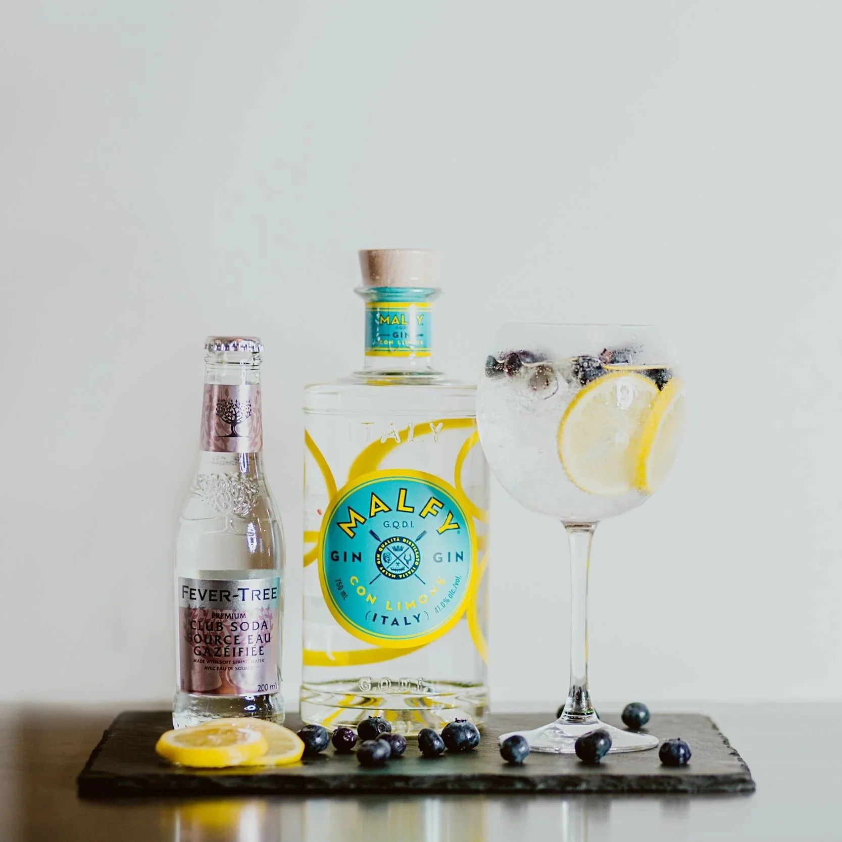 A bottle of Malfy gin with lemon flavor, a bottle of Fever-Tree tonic water, and a large glass of gin and tonic garnished with lemon slices, blueberries, and blackberries, arranged on a wooden surface against a plain background.