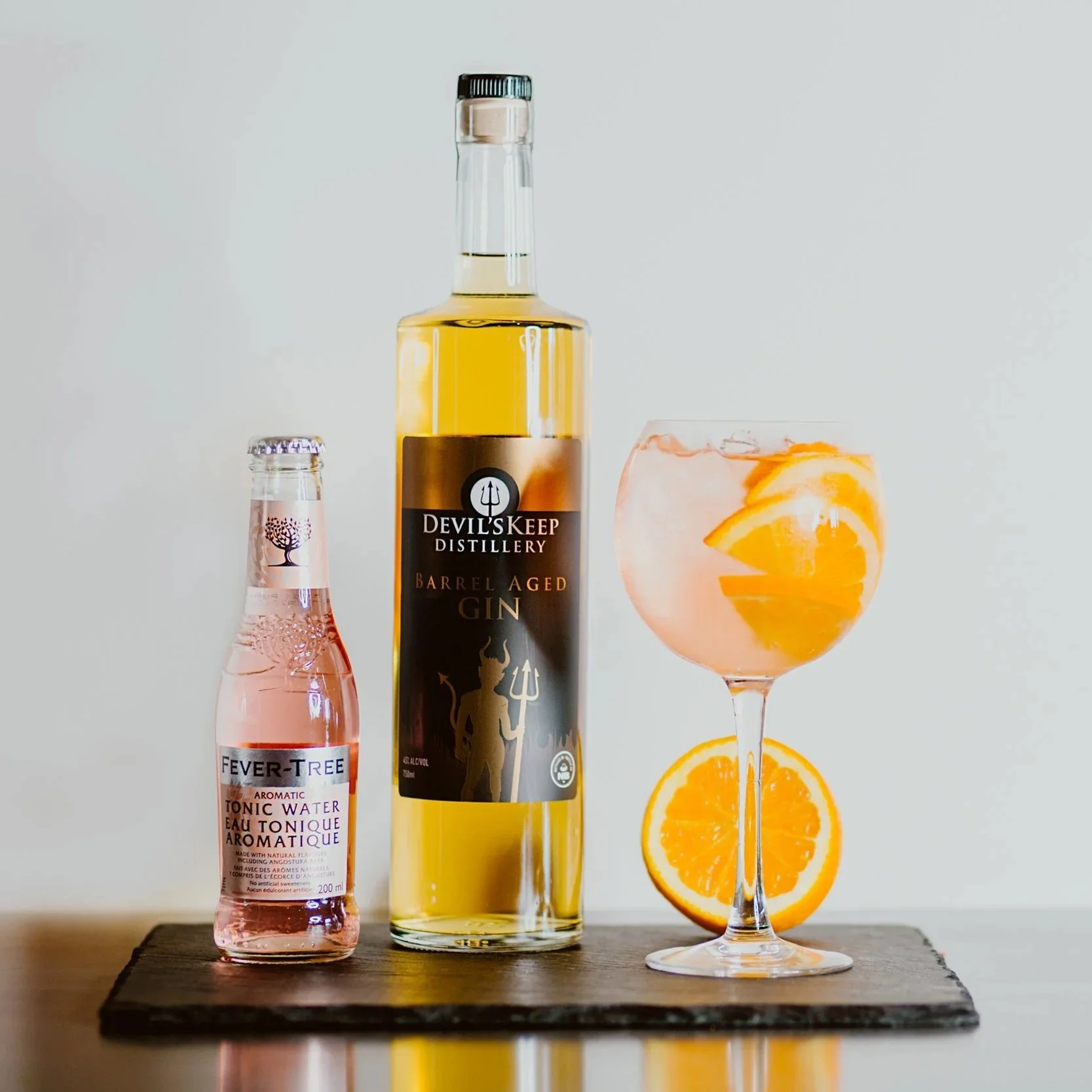 A bottle of Devil's Keep Barrel Aged Gin, a small bottle of Fever-Tree aromatic tonic water, and a cocktail glass with orange slices, all placed on a black wooden board against a plain light background.