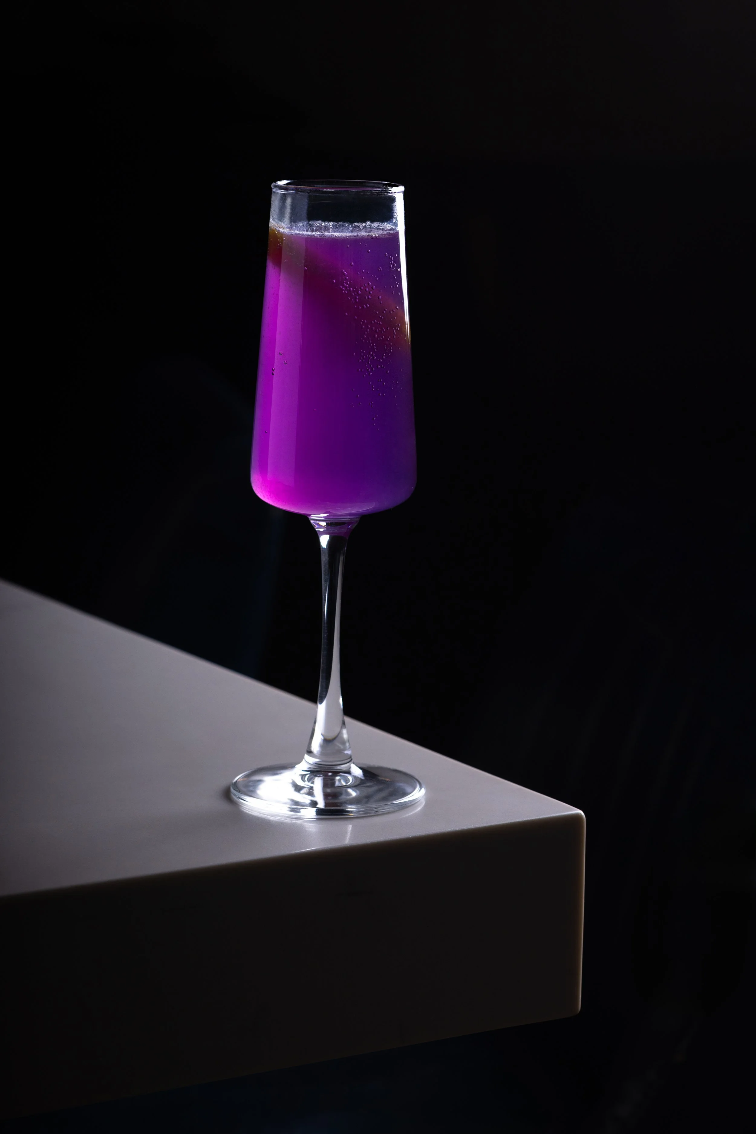A tall champagne glass filled with a purple gin cocktail, Royal Flute, placed on the white bar top of Southside Shake with a black background.