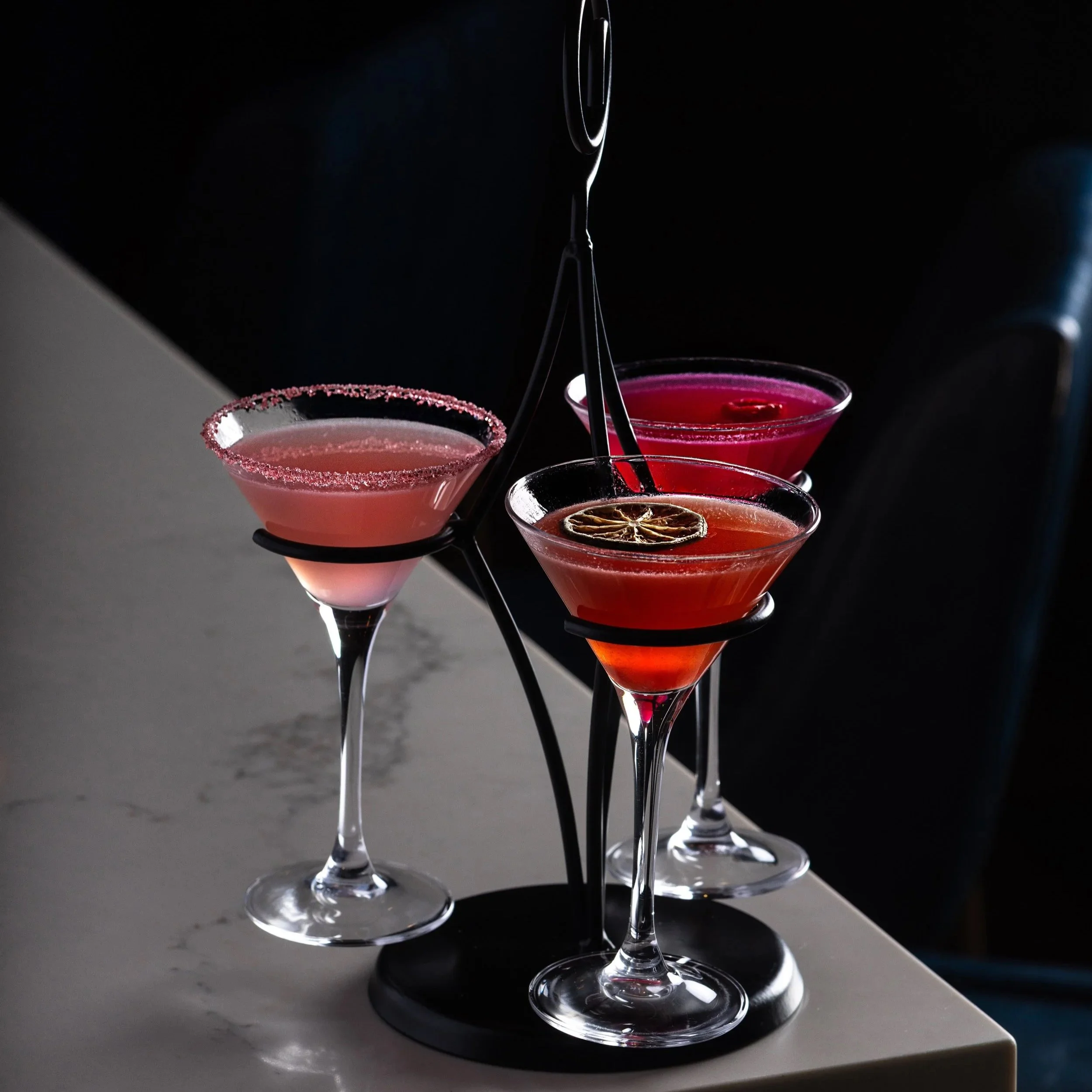 Three colourful Tiny Tini cocktails in martini glasses on a black metal stand, with a dark background and white marble countertop of Southside Shake's bar top.