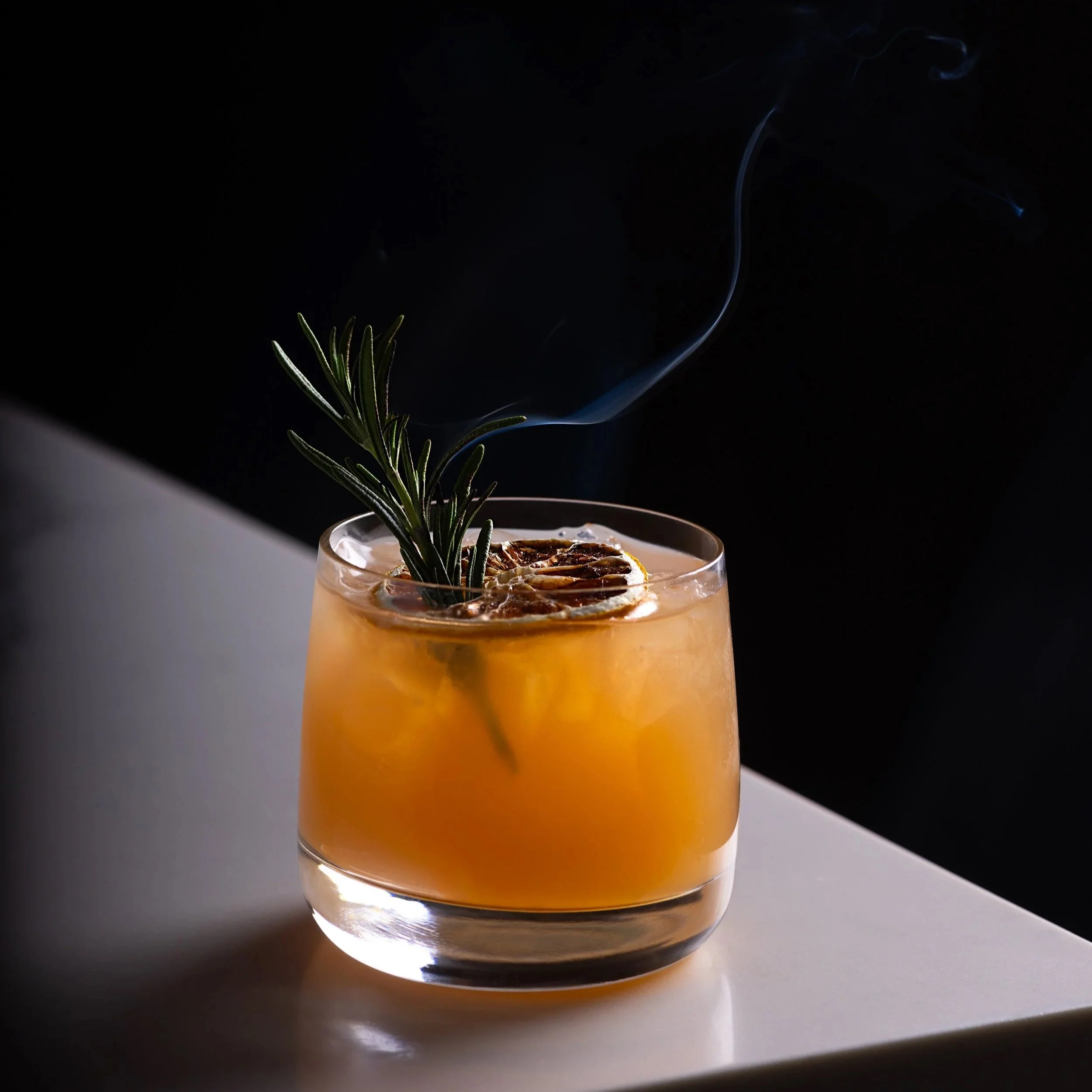 A gin cocktail garnished with a sprig of rosemary and slices of dried citrus, with smoke rising from it, against a dark background at Southside Shake.