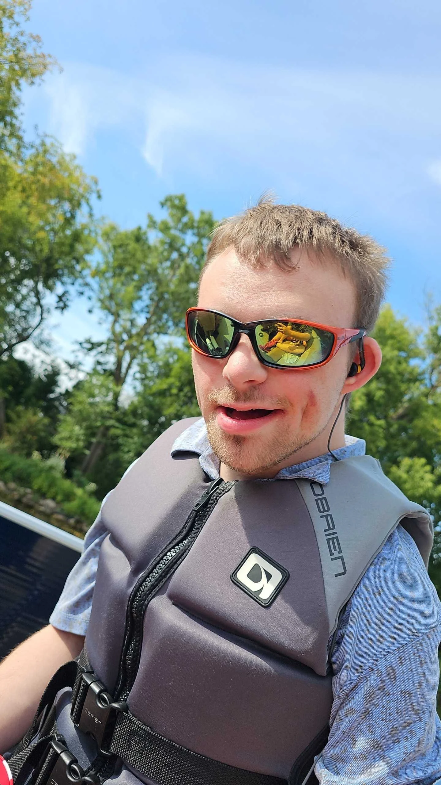 A man wearing sunglasses, a life jacket, and a patterned shirt outdoors on a sunny day surrounded by green trees.