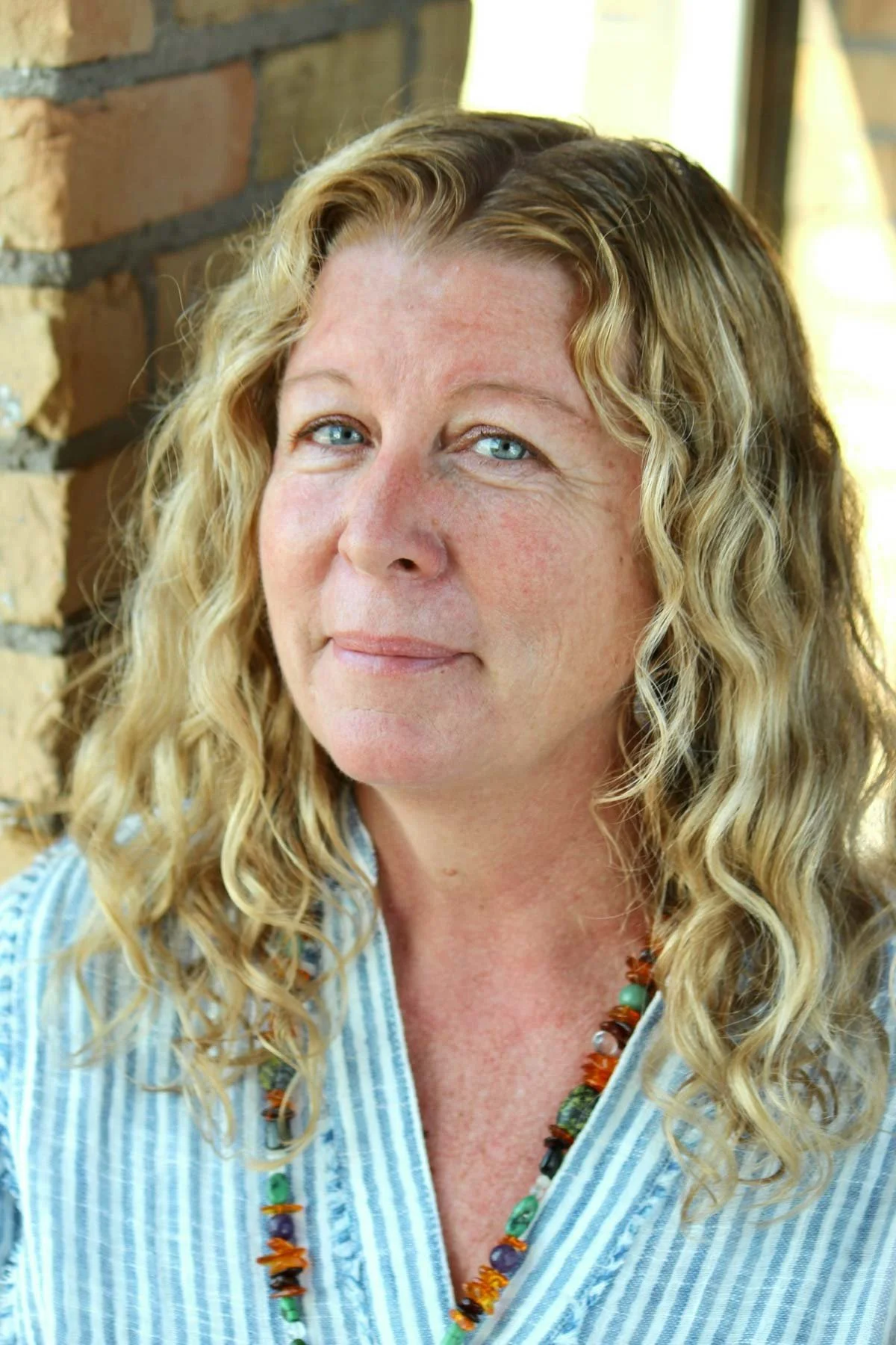 Close-up of a woman with curly blonde hair, blue eyes, wearing a striped shirt and a colorful beaded necklace, standing near a brick wall background.