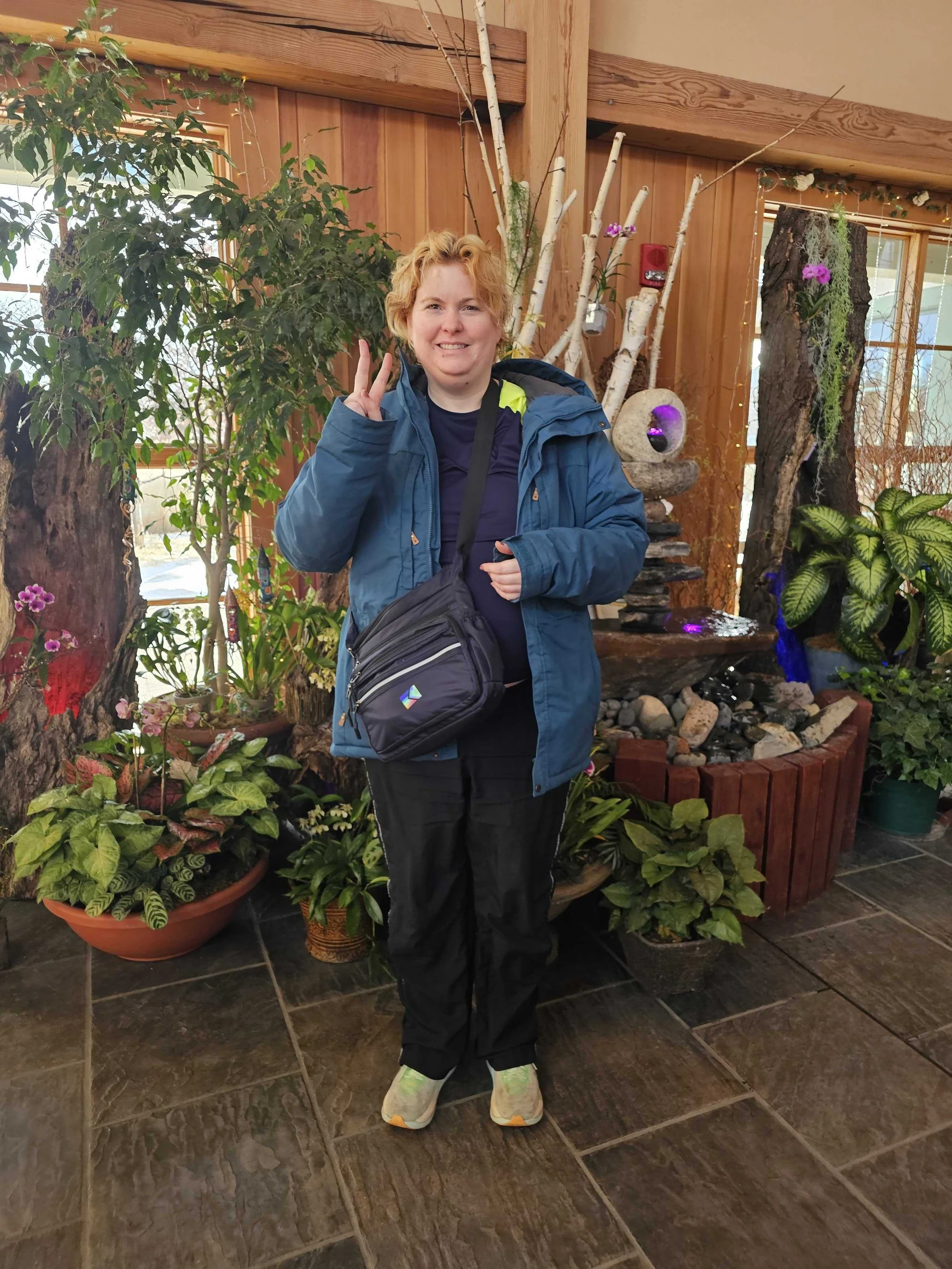 Woman with short blonde hair smiling and making a peace sign, standing indoors in front of lush potted plants and a wooden wall, dressed in outdoor winter clothing.