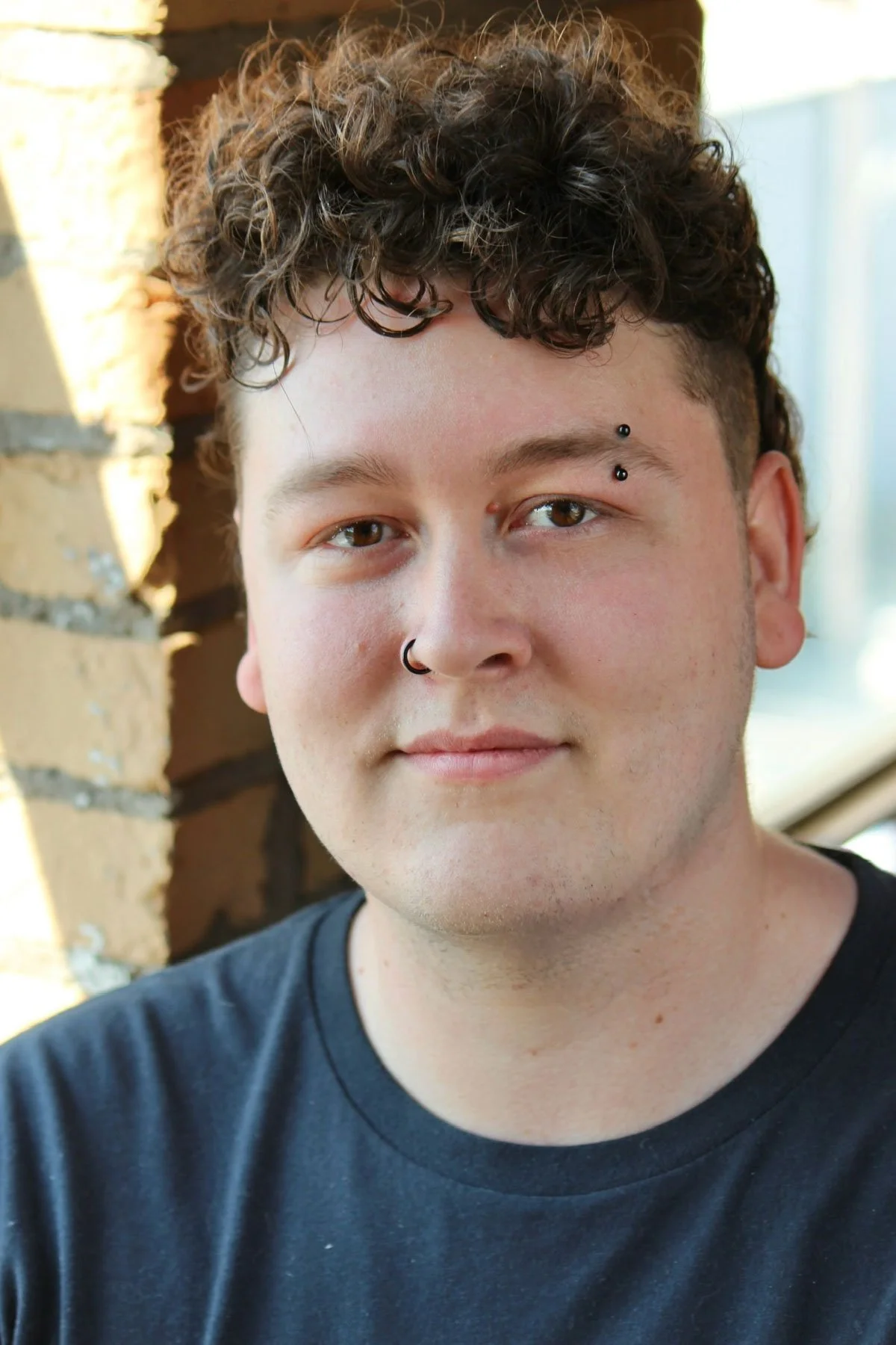 Close-up of a young person with curly brown hair, warm skin tone, and facial piercings, wearing a black t-shirt, standing outdoors near a brick wall with sunlight.
