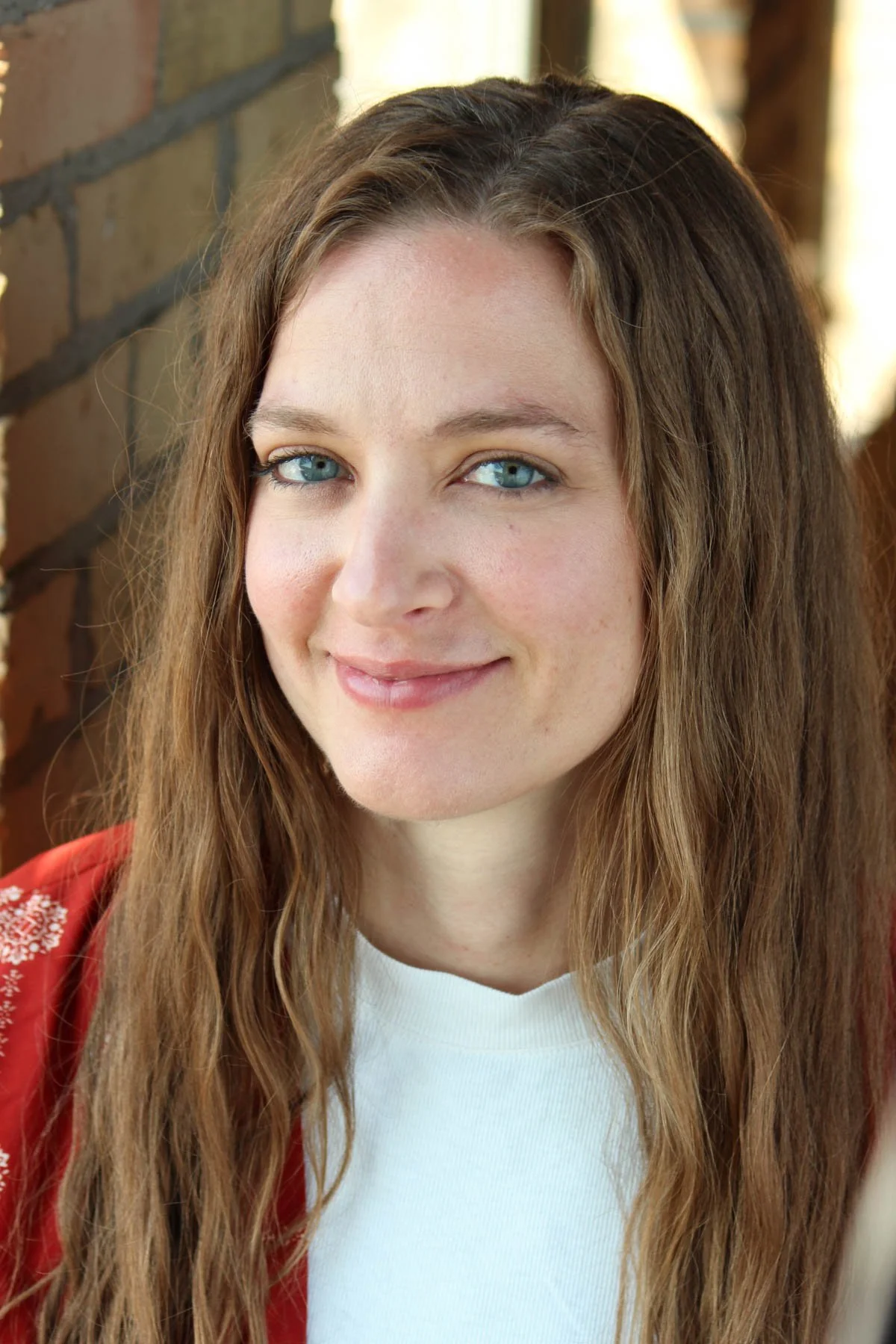 Close-up of a woman with long wavy brown hair, blue eyes, and a fair complexion, smiling softly, wearing a white top and a red jacket, with a brick wall in the background.