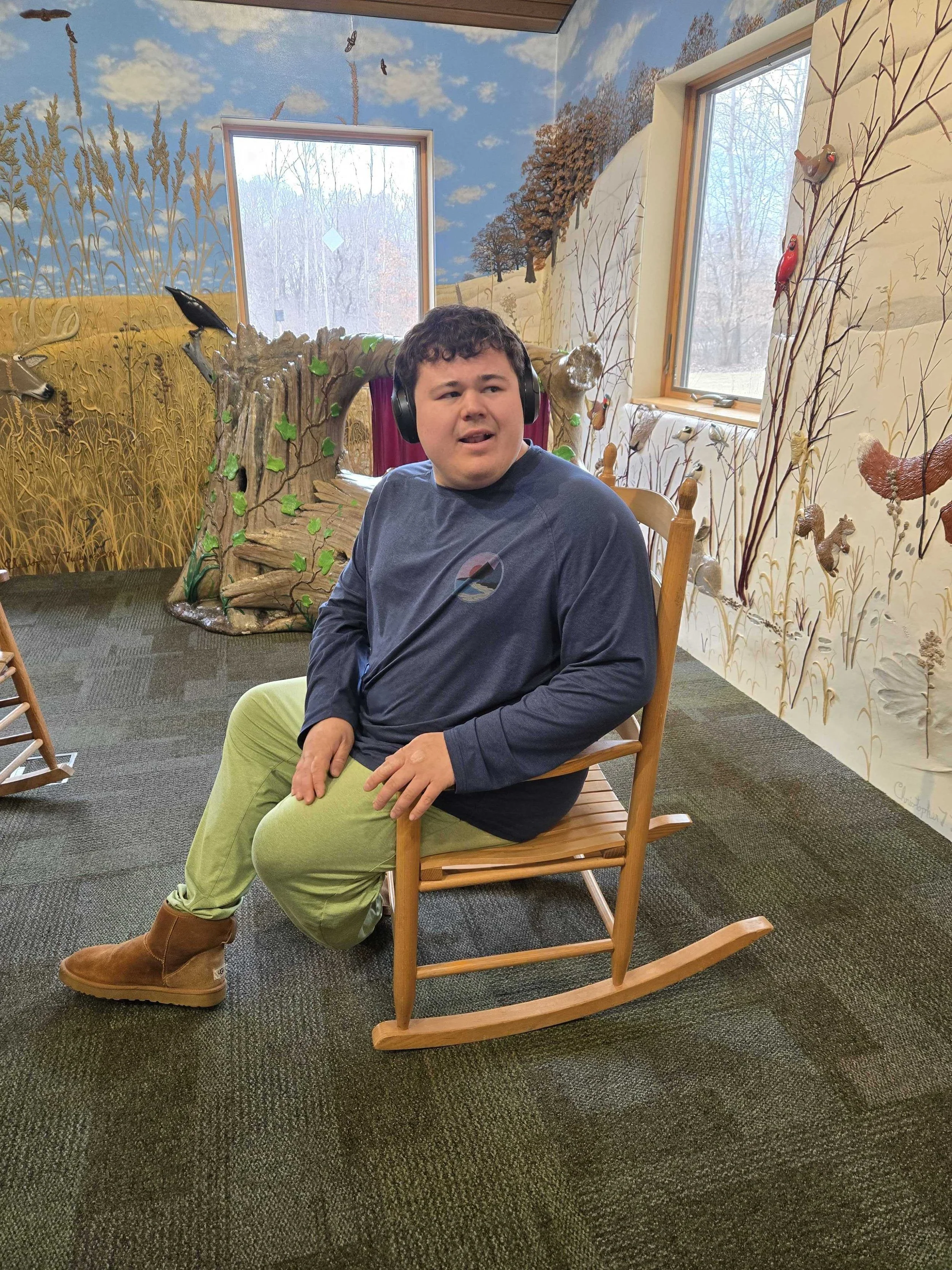 A boy sitting on a wooden rocking chair in a room with nature-themed murals on the walls and windows showing snowy trees outside.