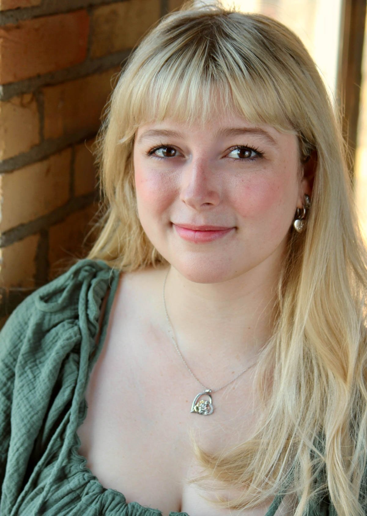 Close-up portrait of a young woman with blonde hair, blue eyes, and a gentle smile, wearing silver jewelry including earrings and a necklace, against a brick wall background.