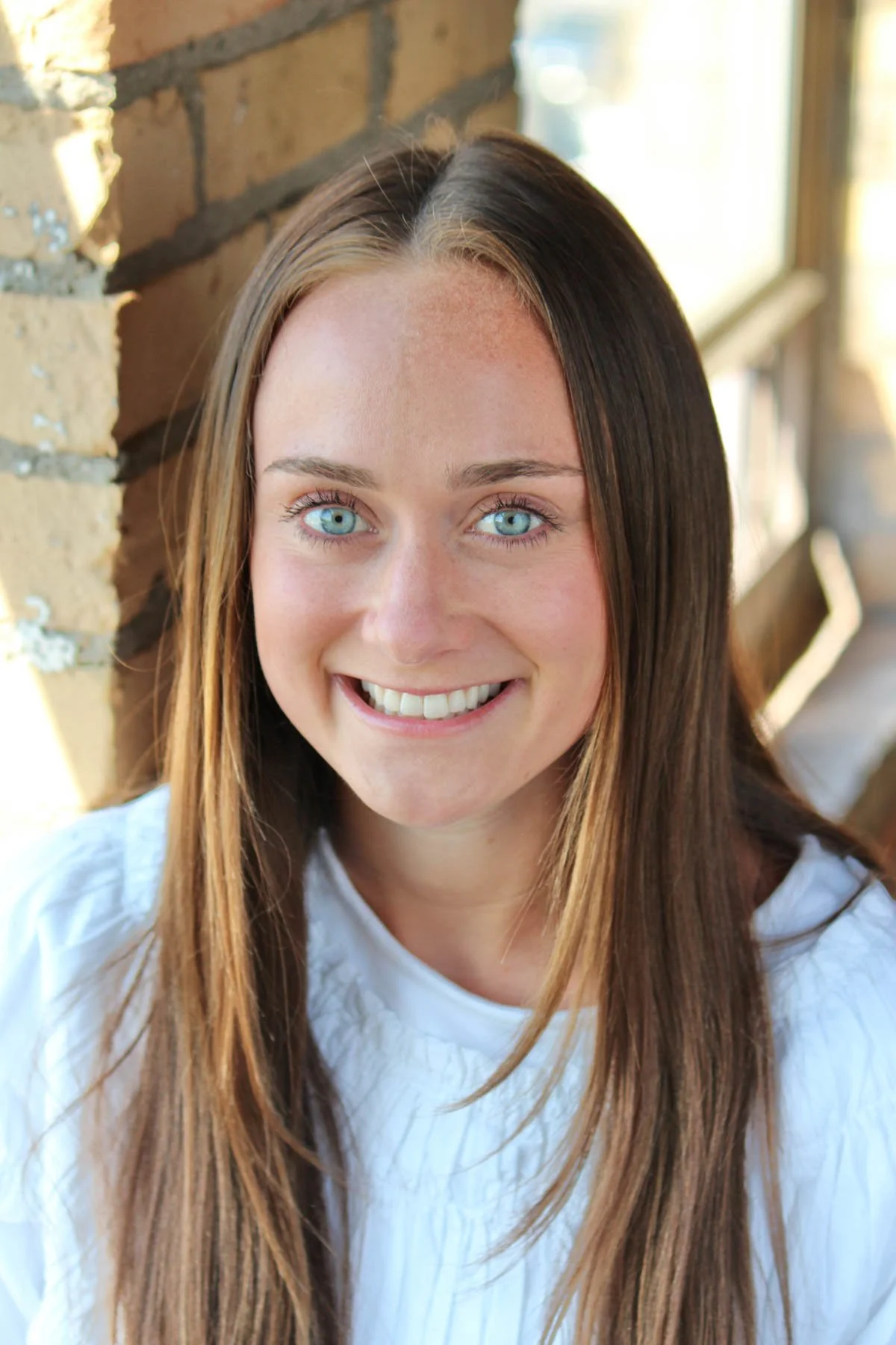 Close-up of a young woman with long brown hair and bright blue eyes smiling at the camera, standing next to a brick wall with sunlight streaming in from the side.