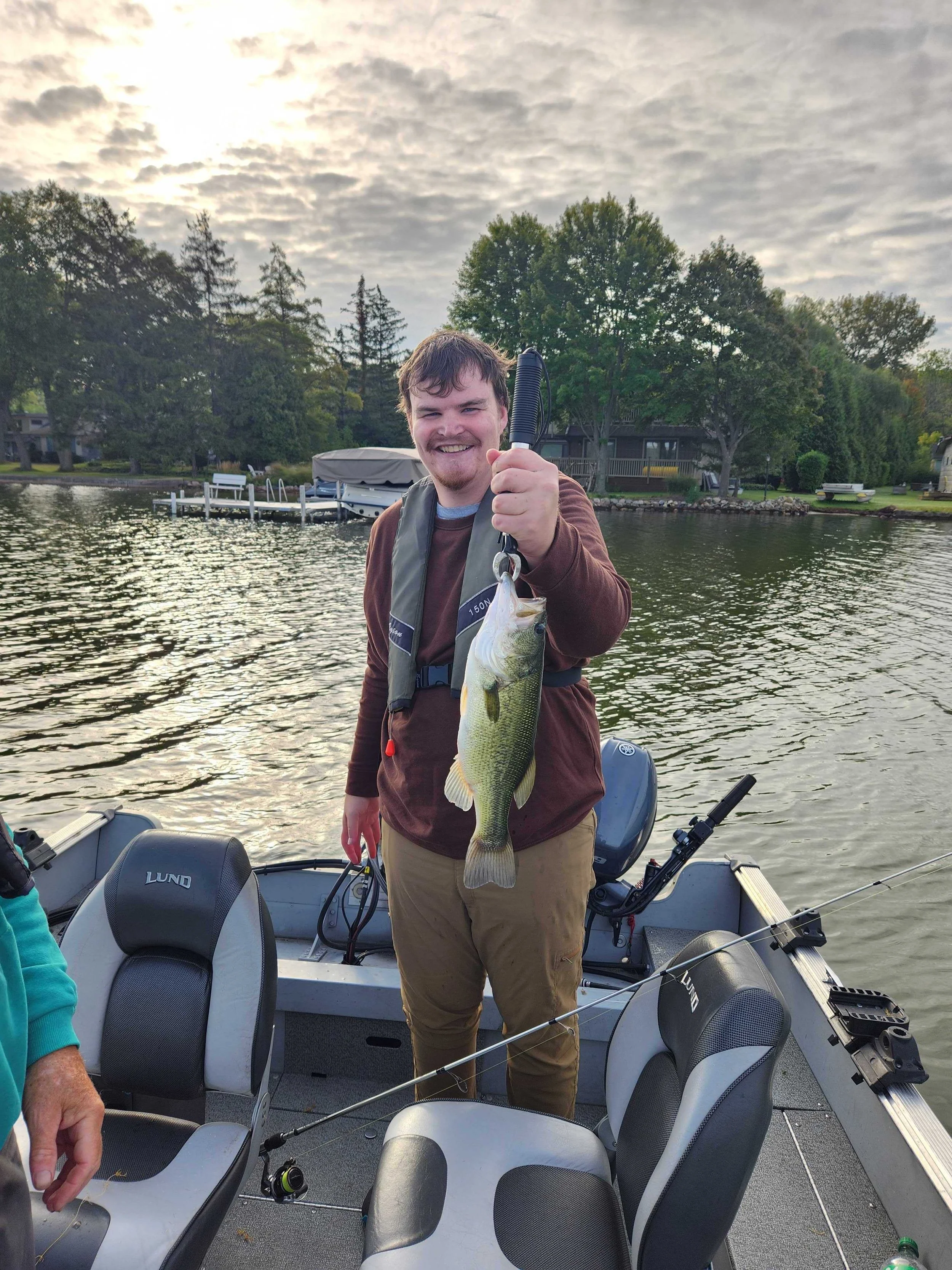 A young man standing on a boat holding a large fish he caught, smiling at the camera, with water and trees in the background during the evening.