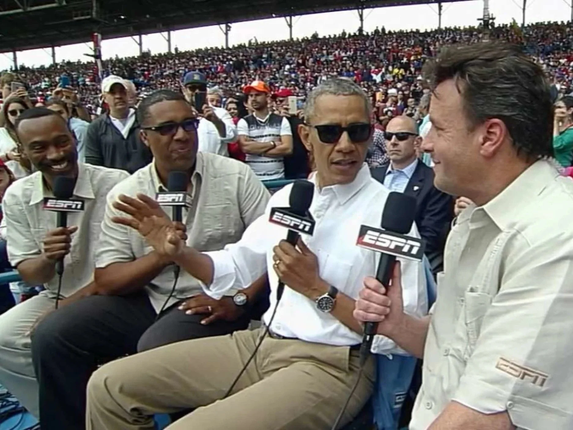 A group of sports commentators holding ESPN microphones at a stadium during a sports event, with a large crowd in the background.