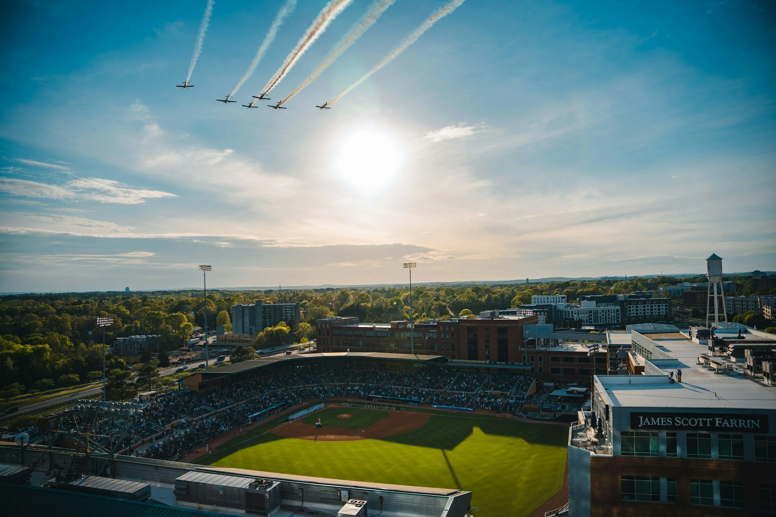 Aerial view of a baseball stadium with the name James Scott Farrin, filled with spectators. The field is under a clear sky, with seven planes flying overhead leaving contrails.