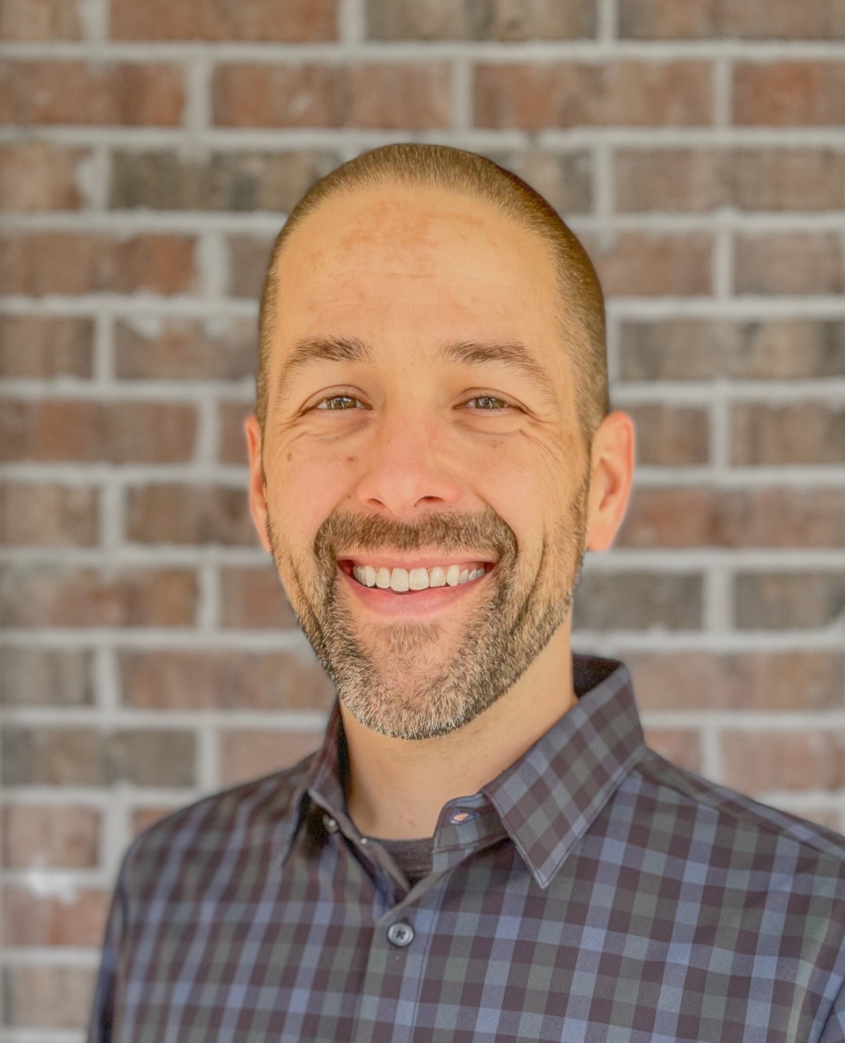 Smiling man with short hair, beard, and mustache wearing a checkered shirt in front of a brick wall.