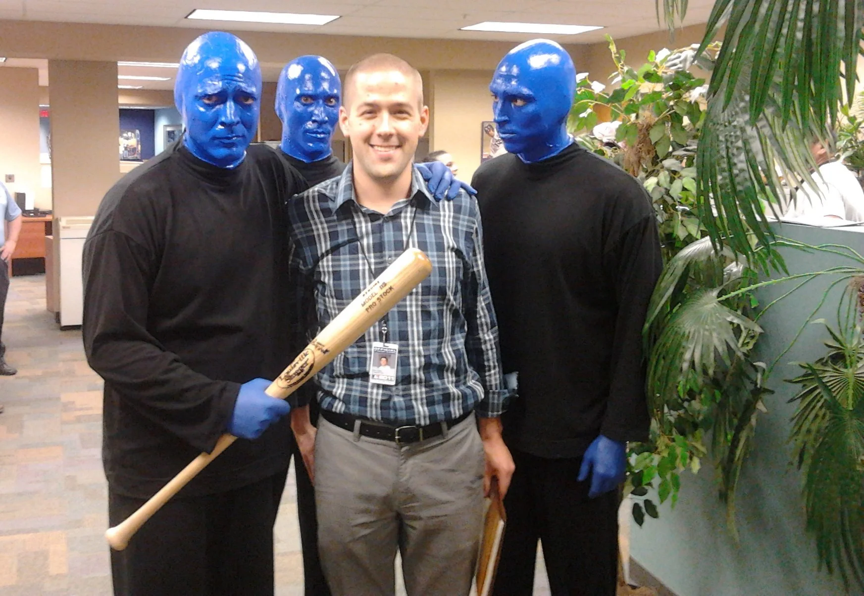 A young man in casual clothing standing between three men dressed in black with blue painted faces, who are wearing gloves and holding a baseball bat and a framed picture, inside a room with potted plants.