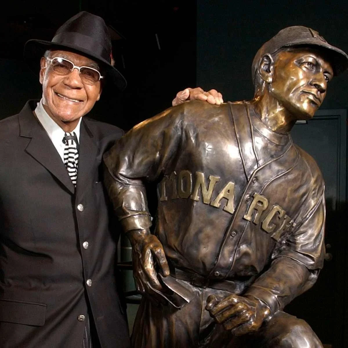 Hall of Famer and Negro Leagues legend Buck O'Neil wearing a black hat, black suit, striped tie and glasses standing next to a bronze statue of himself as a Kansas City Monarchs ballplayer.