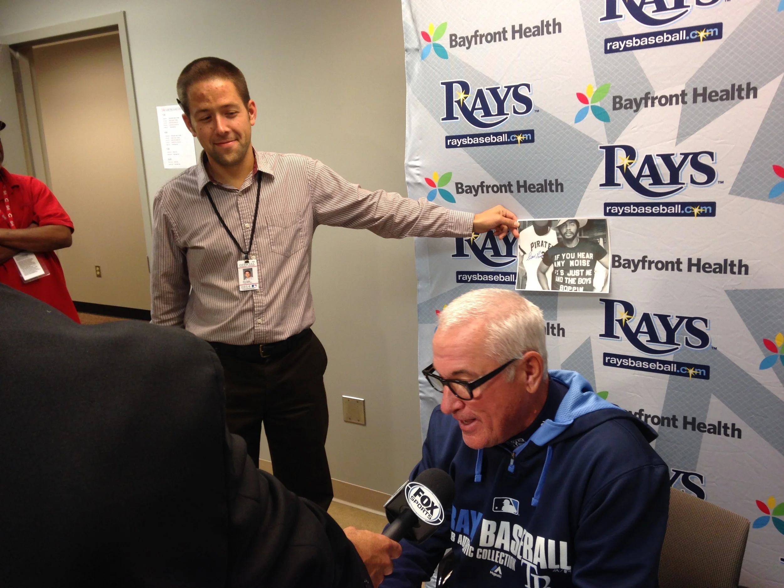 A press conference or interview with a man in a blue Rays hoodie, wearing glasses, sitting in front of a backdrop with Rays and Bayfront Health logos. A reporter holding a microphone with a Fox Sports logo is interviewing him. Another man stands nearby holding a photo, and a person in a red shirt is in the background.