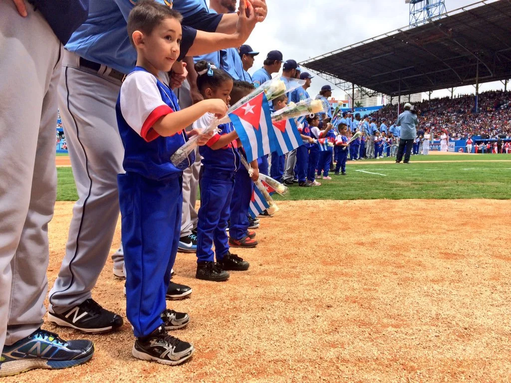 A group of children and adults standing in line on a baseball field, holding flowers and American flags with Cuba flags, during a baseball game or ceremony with a crowd in the stands.