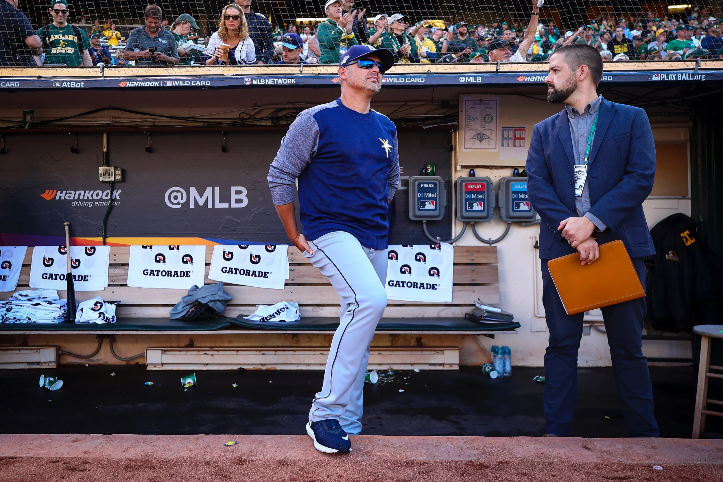 A man in a baseball uniform leaning against a dugout, another man in a suit holding a briefcase, and a crowd watching a baseball game in the background.