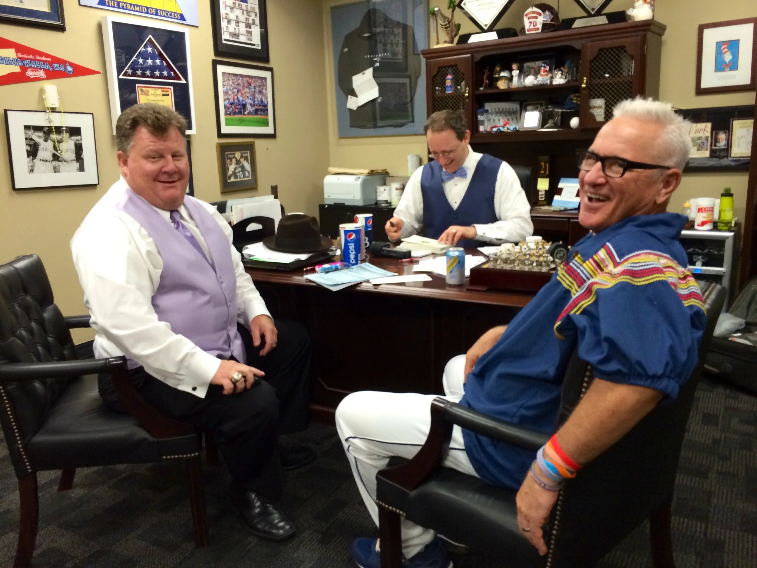 Three men in a festive office, with framed sports memorabilia on the wall. Two men are laughing, one dressed in a purple vest and white shirt, and the other in a blue jacket with colorful stripes, both sitting in chairs. The third man, wearing a vest and bow tie, is seated at a desk with papers, drinks, and office supplies.