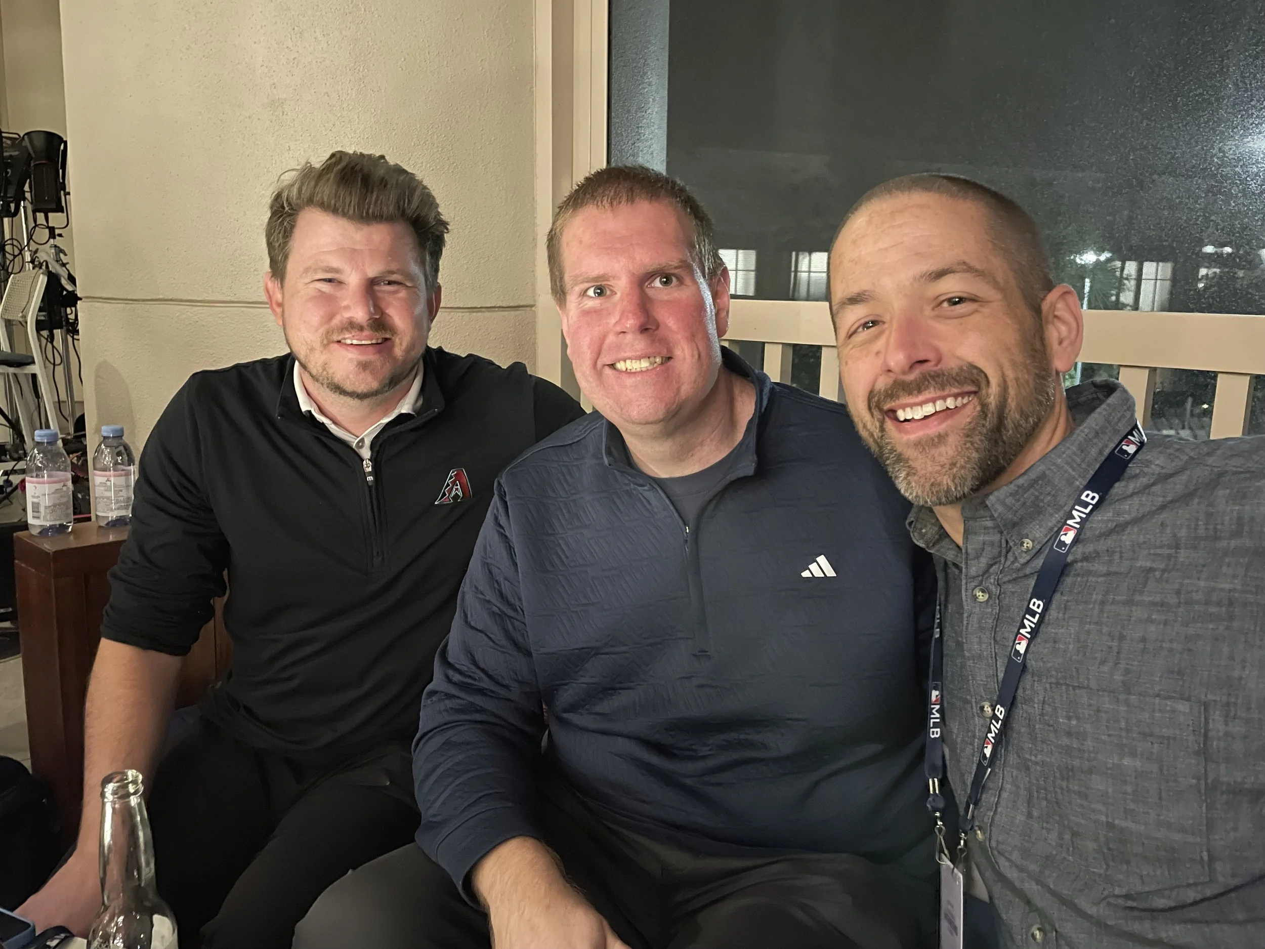 Three men smiling and sitting close together at a table in an indoor setting, with water bottles in the background and one man wearing an MLB lanyard.