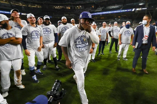 Baseball players and staff celebrating on the field after a game, with some wearing 'American League' shirts and hats, and a player at the center laughing and covering his face.