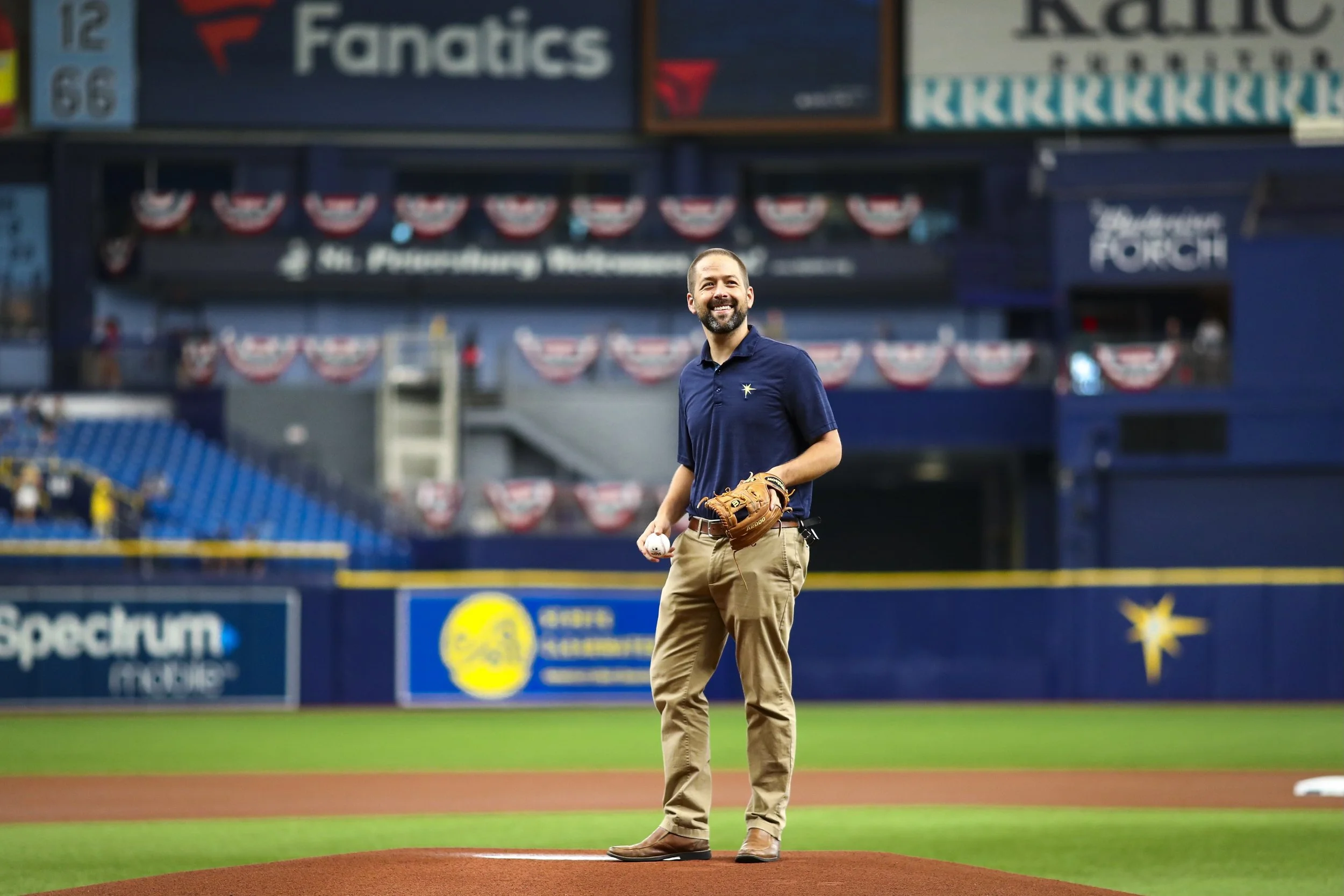 A man standing on a baseball field holding a baseball and wearing a baseball glove, smiling, with stadium seating and banners in the background.