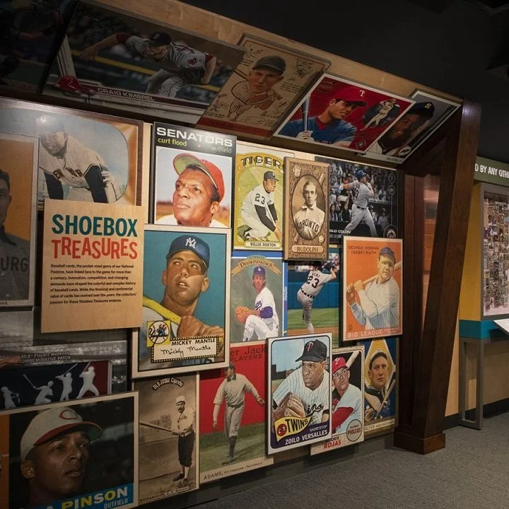 Collection of vintage baseball and sports trading cards and photographs displayed on a wall and a shelf in a sports museum or collectible exhibit.
