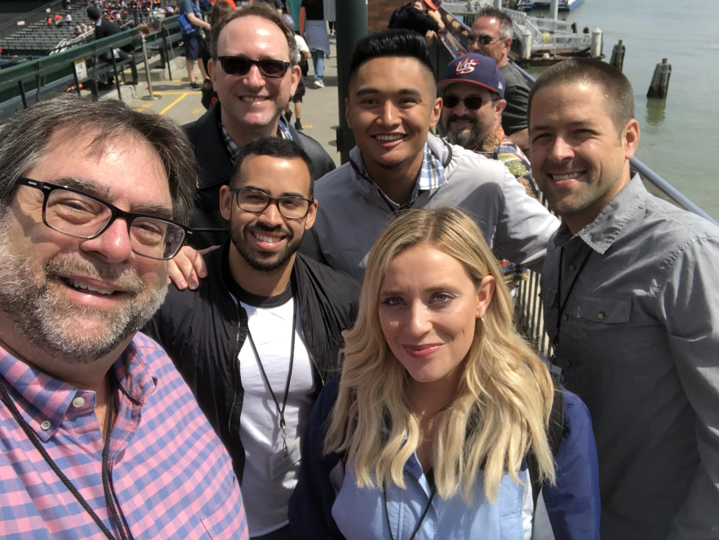 Group of eight people taking a selfie outdoors near water, smiling, with some wearing sunglasses and casual clothing.