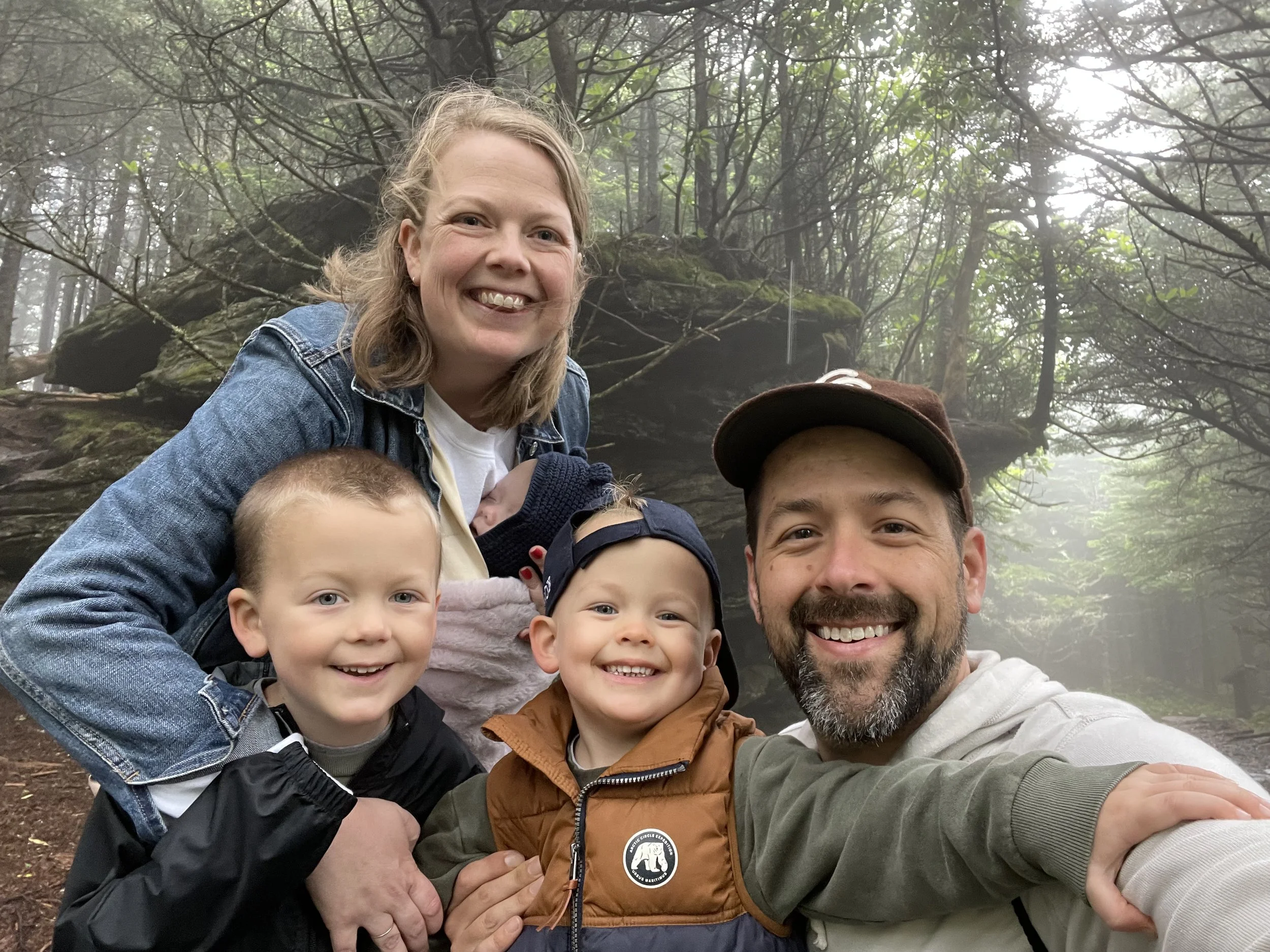 A family of five taking a selfie in the woods on a foggy day. The family includes a woman, a man, and three young children, all smiling at the camera.