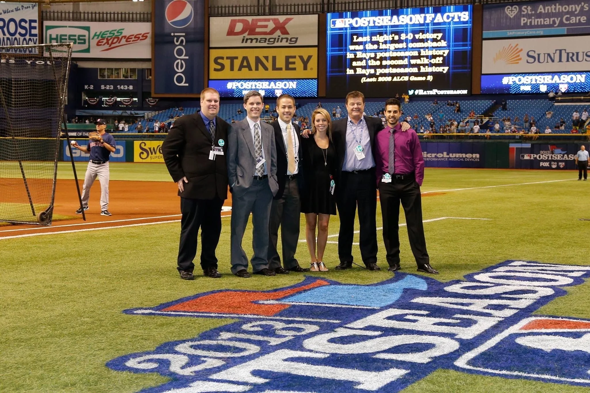 Group of six people dressed in formal attire standing on a baseball field at a stadium, with a large '