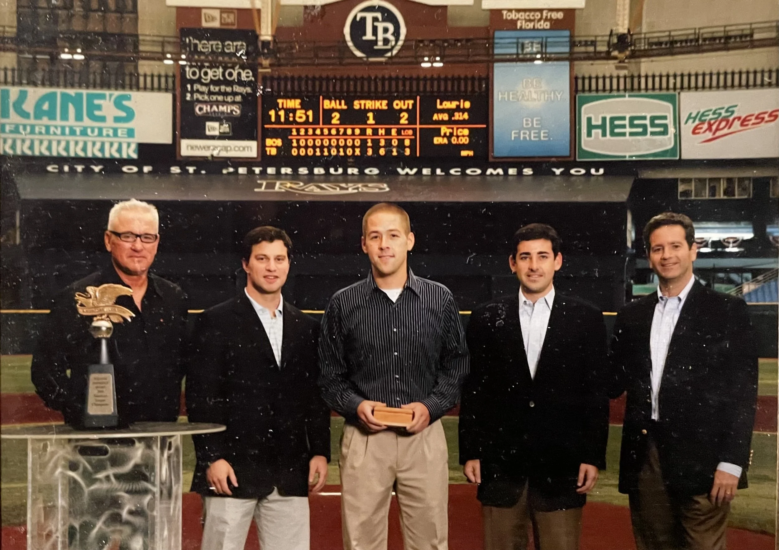 Group of five men standing on a baseball field at PNC Park in Pittsburgh, Pennsylvania, celebrating an award ceremony with a trophy on a stand in front of them. The background shows the scoreboard and advertisements.