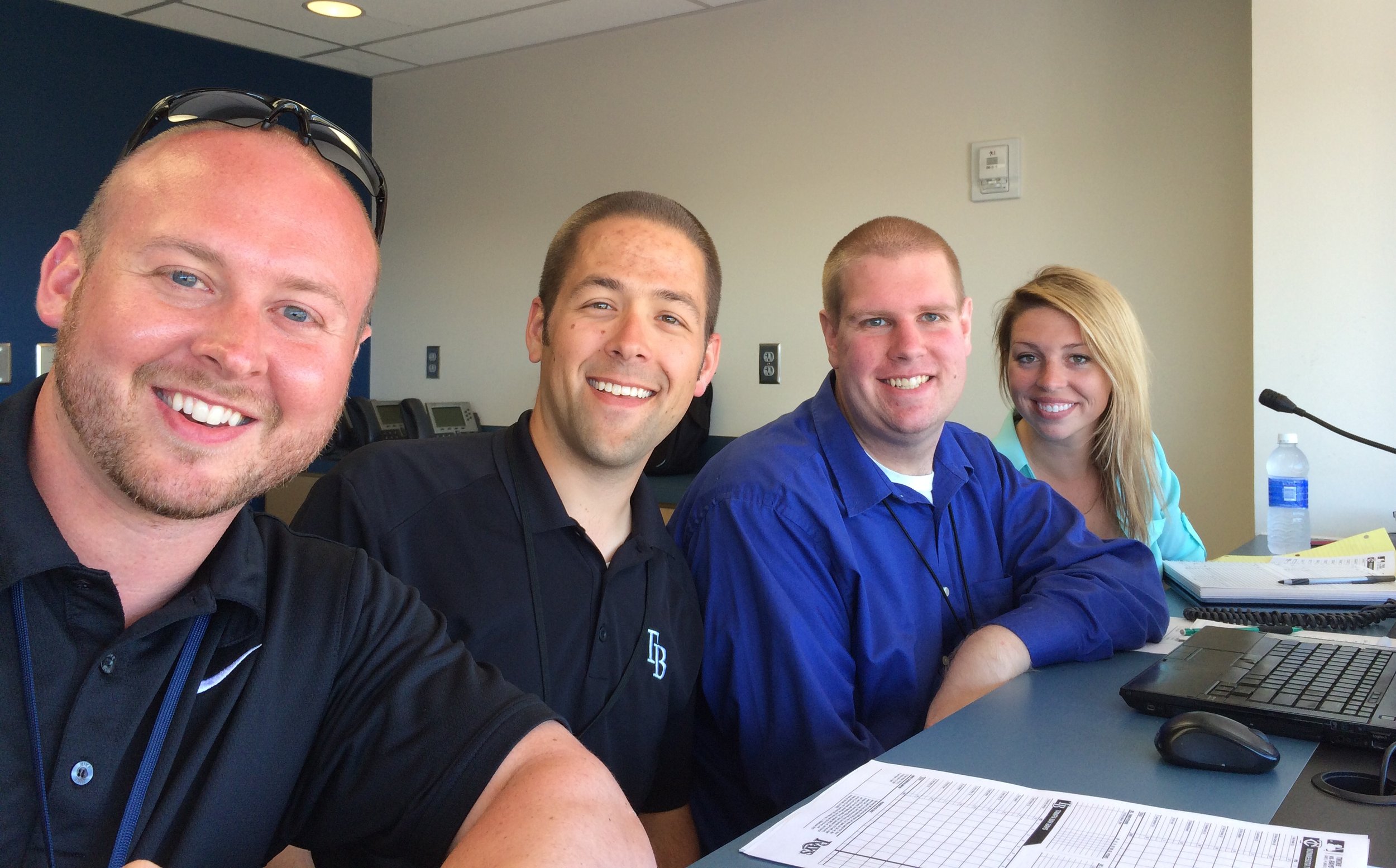 Four people sitting at a table in an office, smiling at the camera, with office supplies, a laptop, a bottle of water, and documents on the table.
