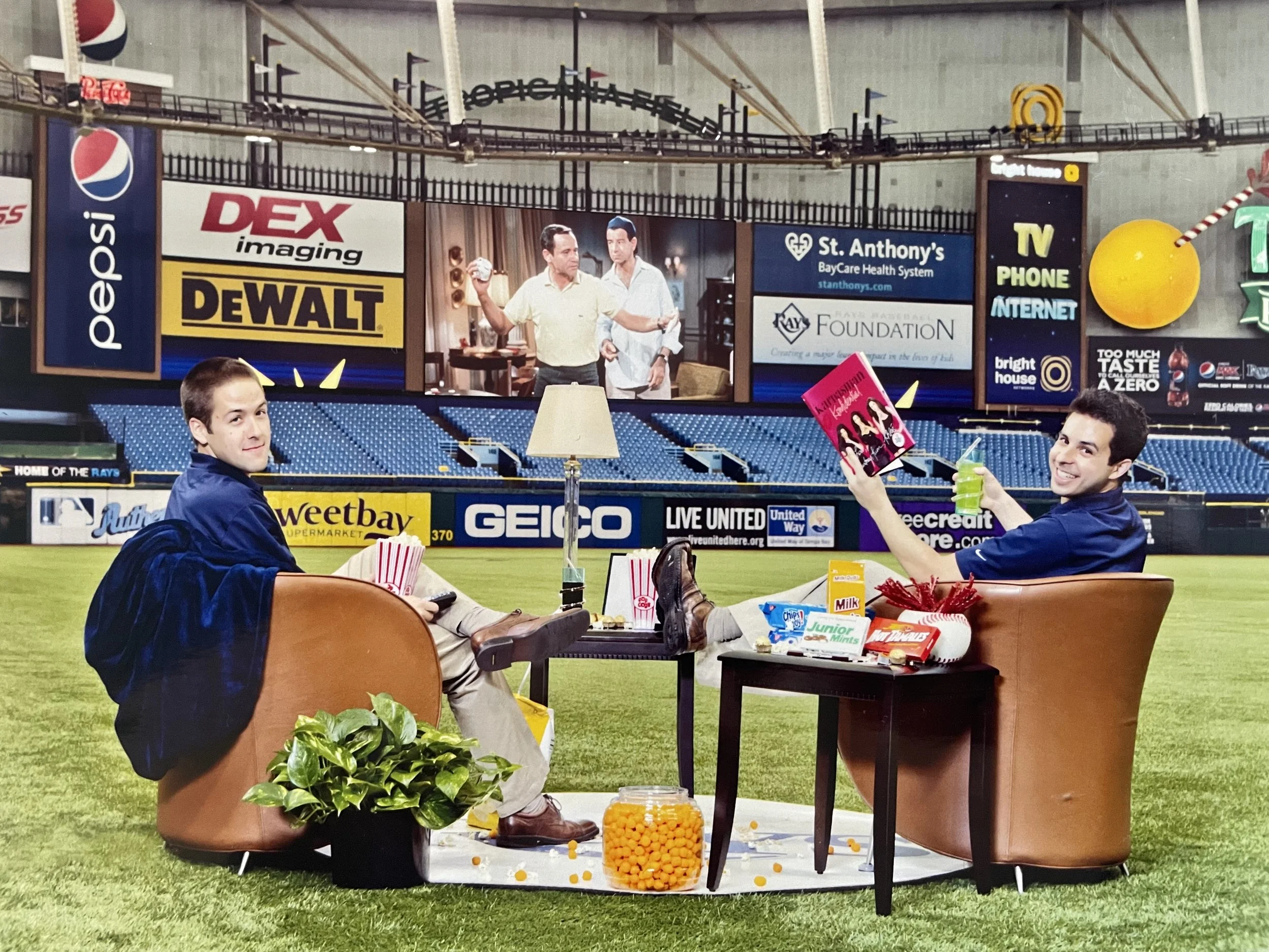 Two young men sitting in brown armchairs on a baseball field, enjoying snacks and drinks, with a small table between them holding popcorn, candy, and drinks. One is holding a book and smiling, the other has a green drink. There is a large digital screen and various advertisements in the background.