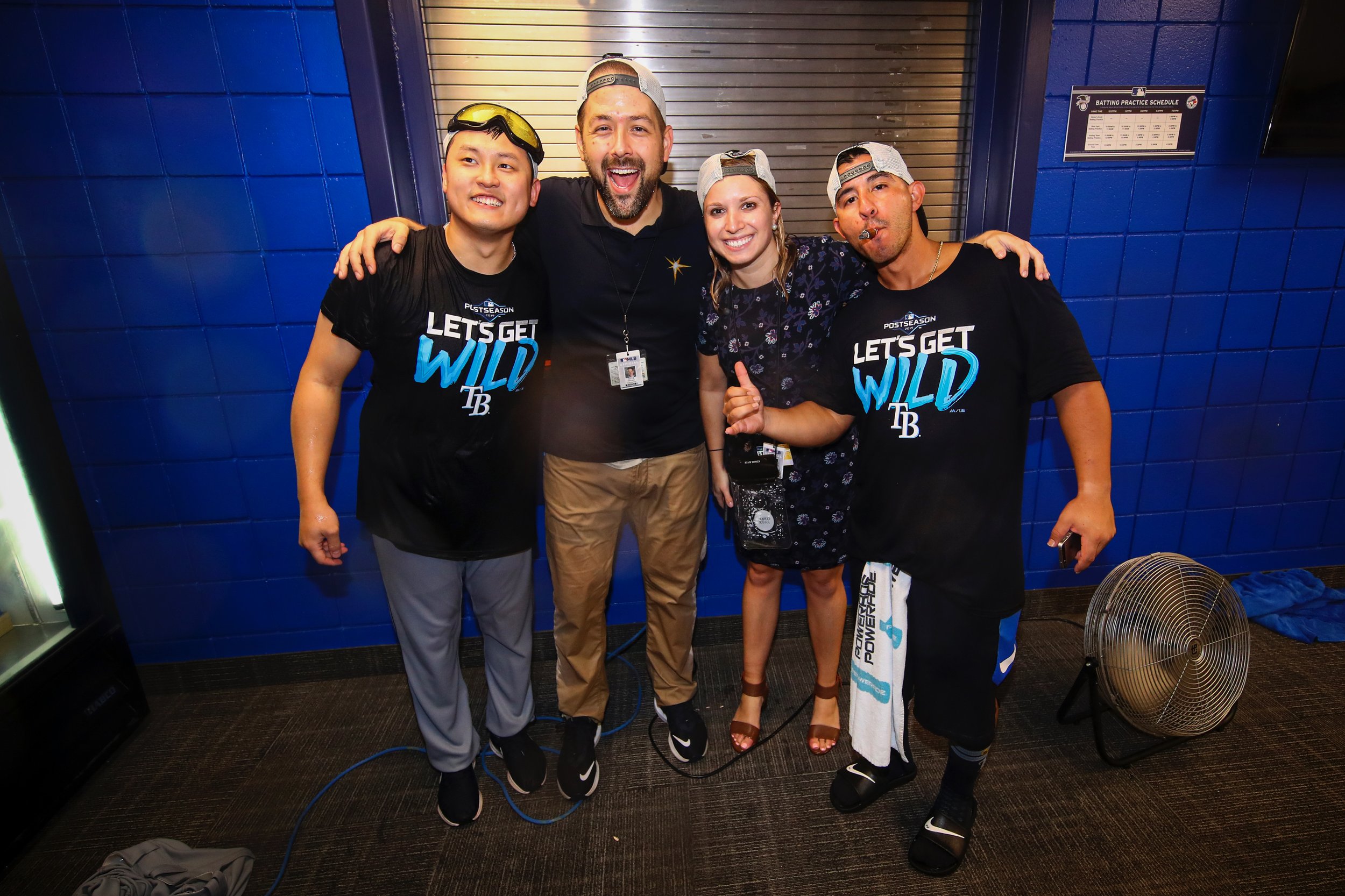 Group of four celebrating baseball victory, two men wearing Tampa Bay Rays T-shirts and two individuals, one man and one woman, dressed casually, standing together inside a sports facility with blue tiled walls and a fan on the floor.