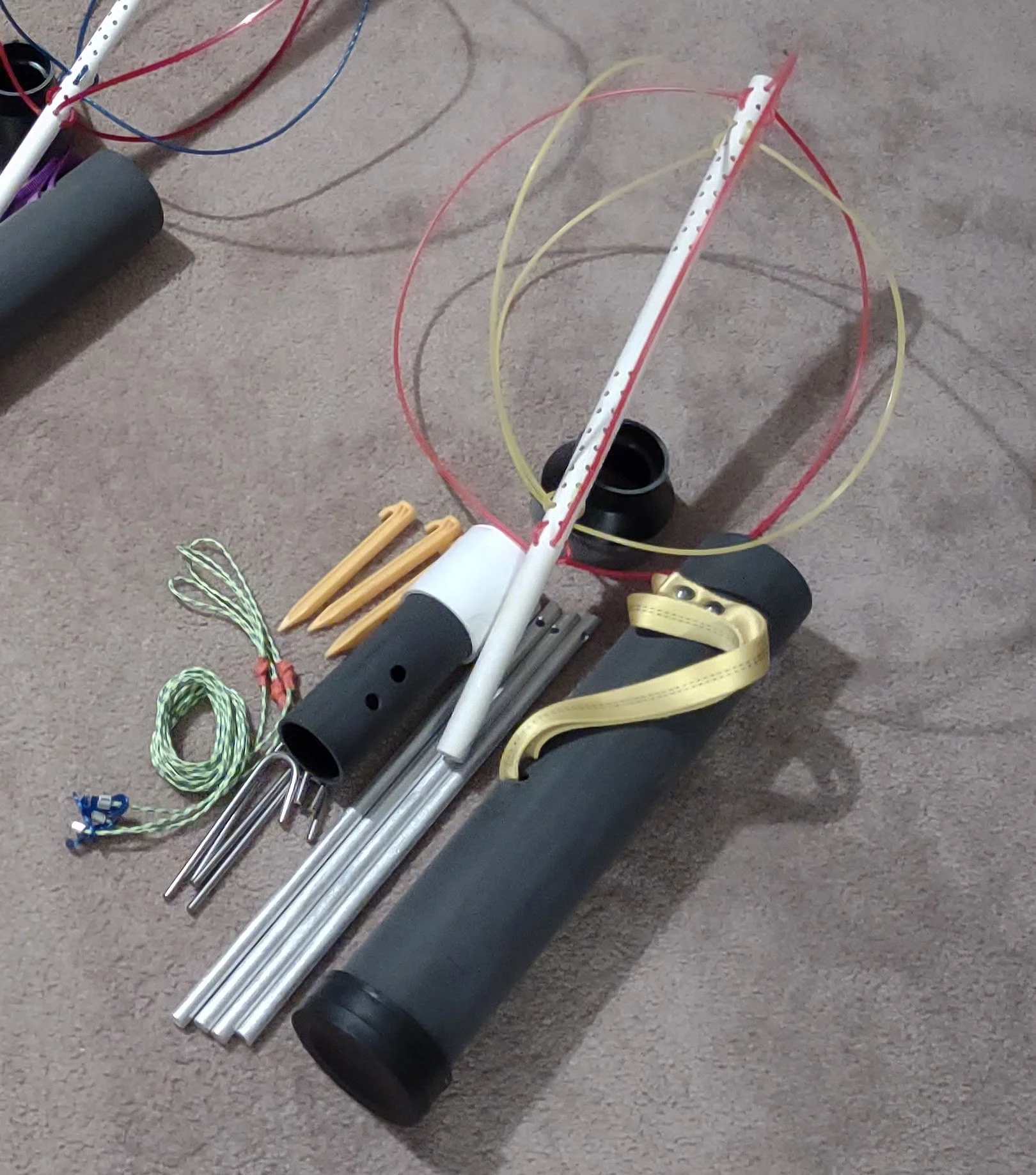 Disassembled jump rope with black foam handles, metal rods, yellow and green ropes, a white plastic stretch, and small yellow plastic components on a carpeted floor.