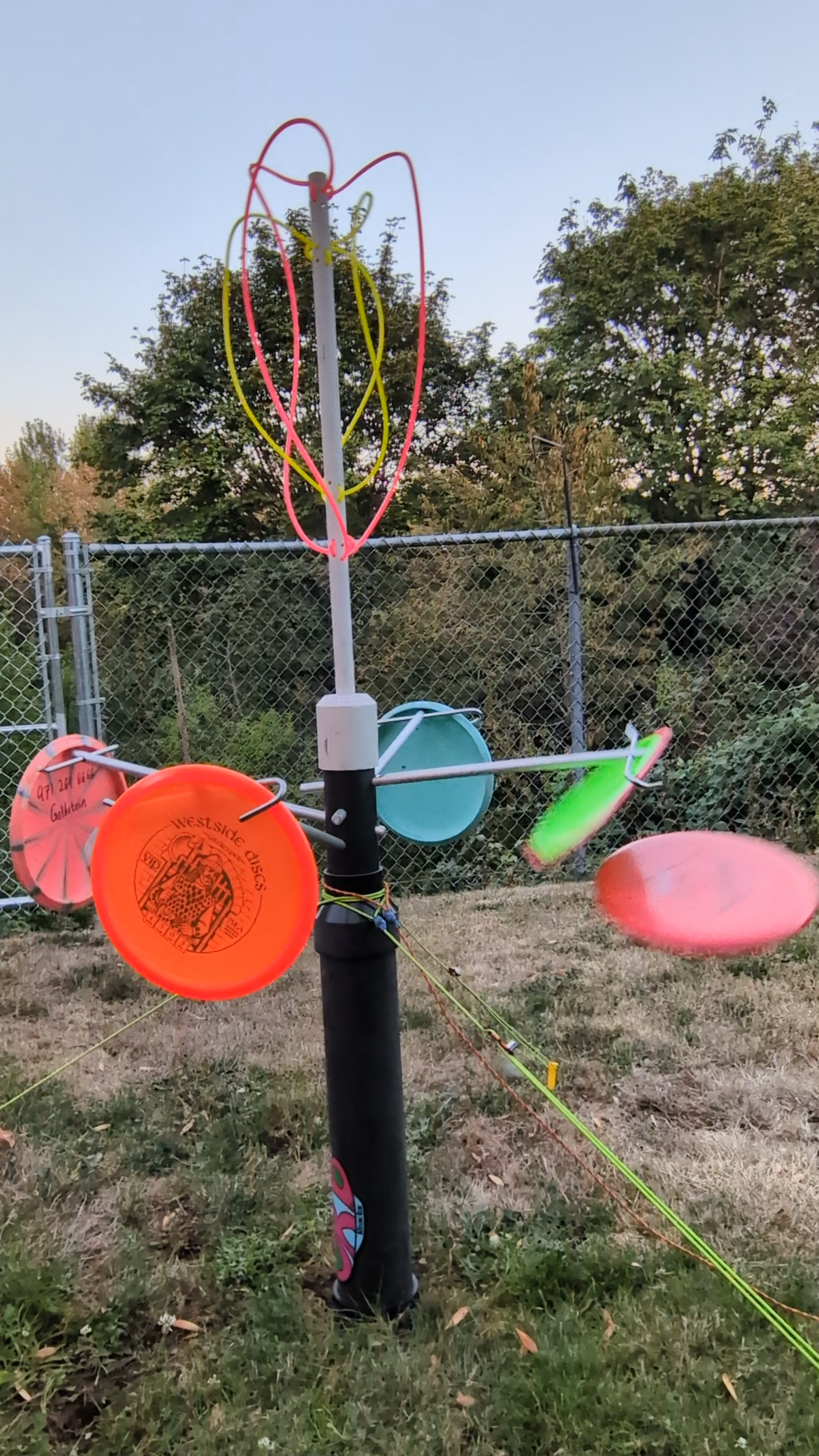 An outdoor kinetic sculpture made of colorful frisbees, sticks, and string mounted on a black pole, set against a fenced yard with trees.