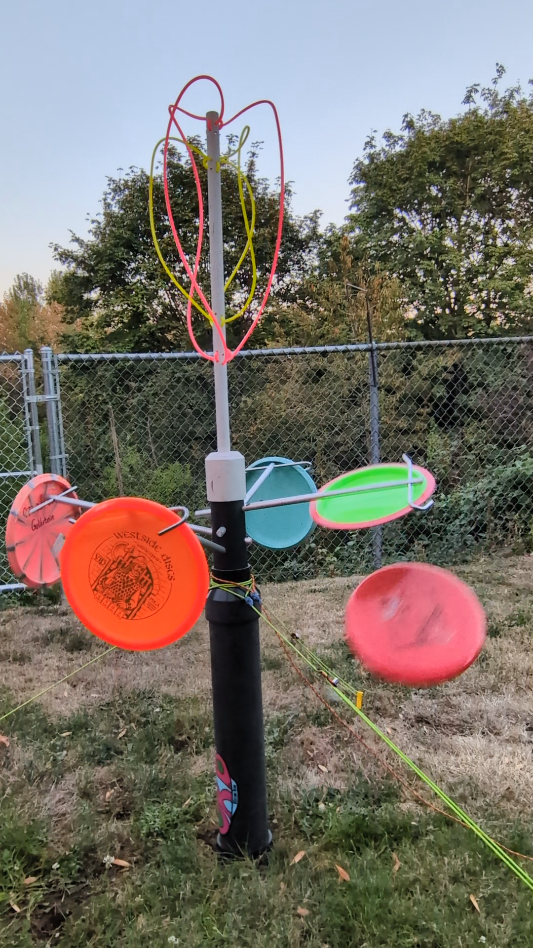 A colorful tree-shaped disc golf target with multiple flying discs attached to it, set outdoors near a chain-link fence and trees.