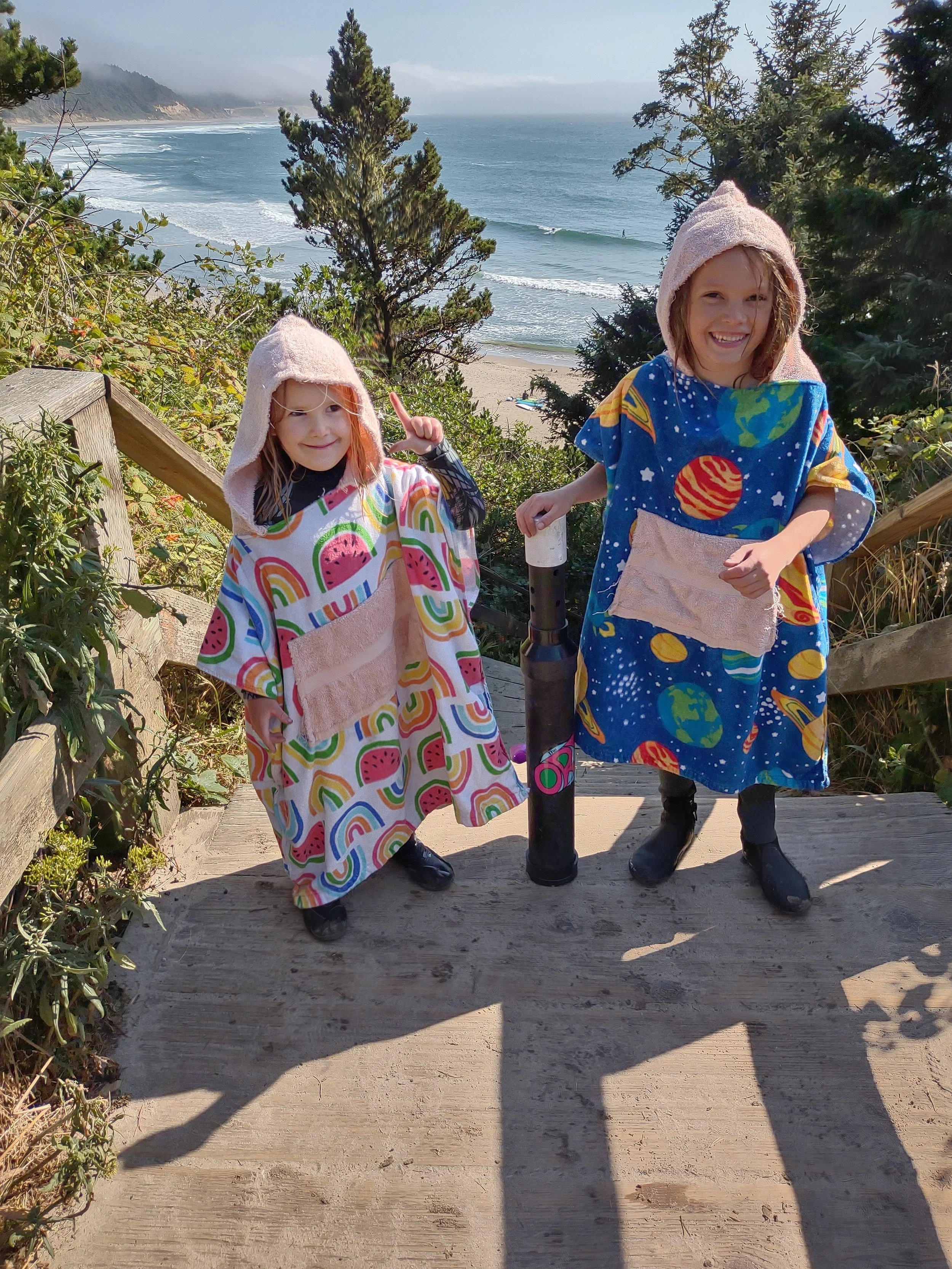 Two children in colorful, space-themed ponchos and hooded towels on a wooden staircase leading to a beach with ocean waves, sandy shore, trees, and distant cliffs.