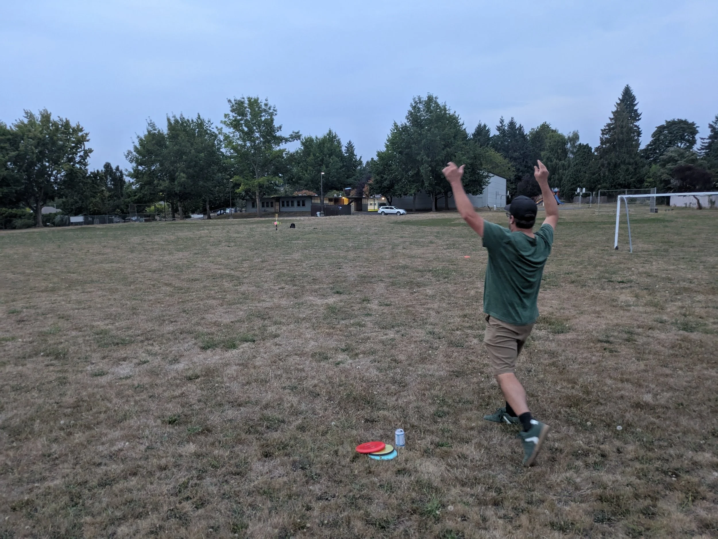 A man in a green shirt and khaki shorts is playing disc golf on a grassy field during early evening, with trees and houses in the background, and disc golf discs on the ground.