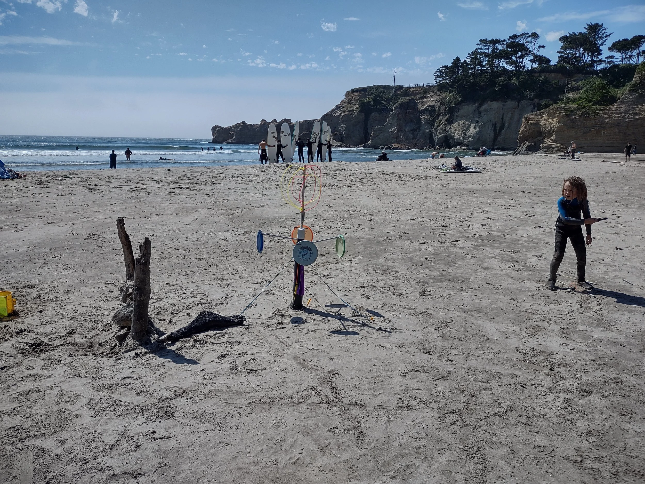 A sandy beach with people in the distance near the water, some standing with surfboards. There is a child with curly hair on the right holding an object, and a small colorful tower with rings and discs in the foreground. Cliffs and trees are visible in the background under a partly cloudy sky.