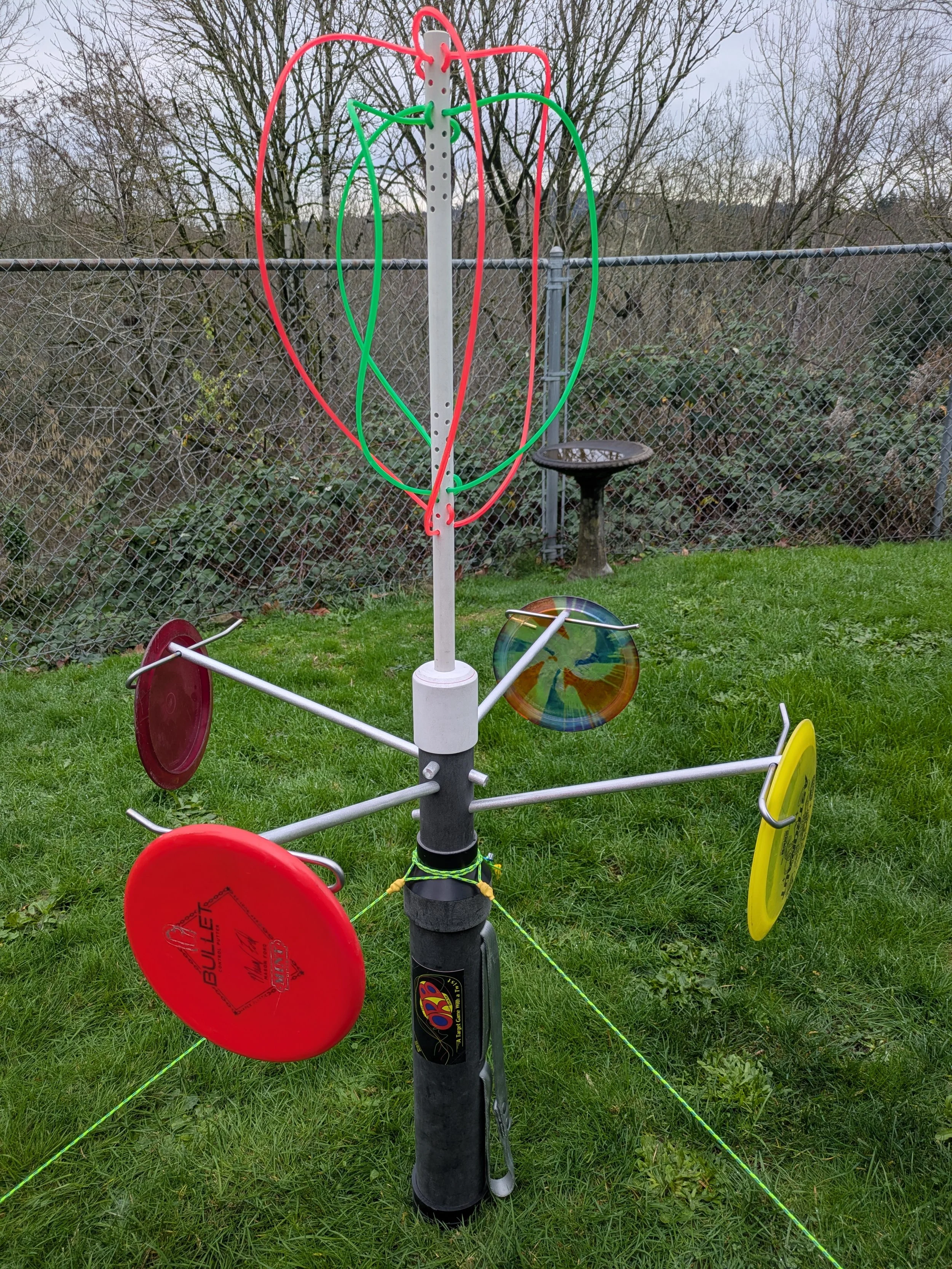 A colorful outdoor game apparatus with various spinning discs and hoops, set on a grassy area enclosed by a chain-link fence with leafless trees in the background.